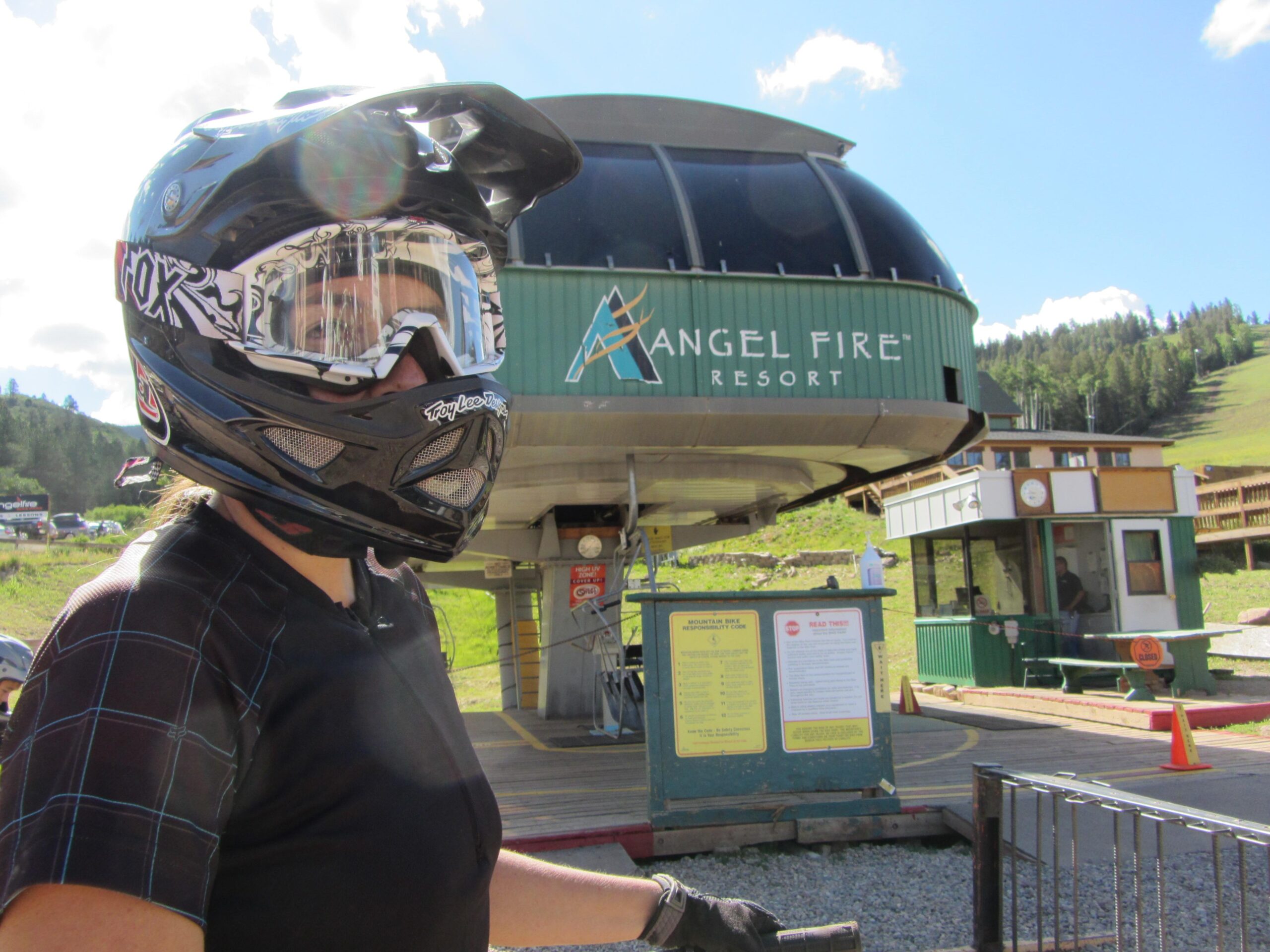 A person wearing a black helmet and goggles stands near a ski lift station at Angel Fire Resort, with green mountains and a clear blue sky in the background. The lift station features a large circular structure above, and various signage is visible nearby. Angel Fire Bike Park mountain bike trail.