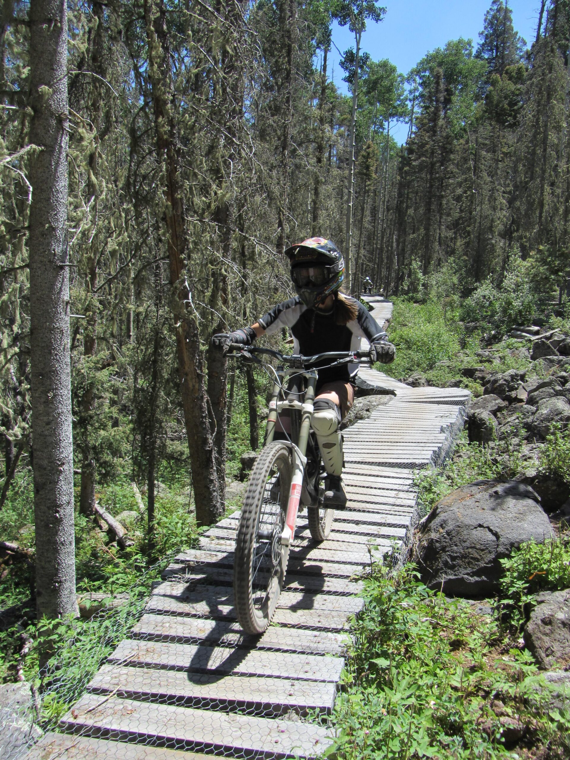 A person riding a mountain bike along a narrow wooden bridge in a forested area. The rider is wearing a helmet and protective gear, navigating through tall trees and greenery under a clear blue sky. Angel Fire Bike Park mountain bike trail.