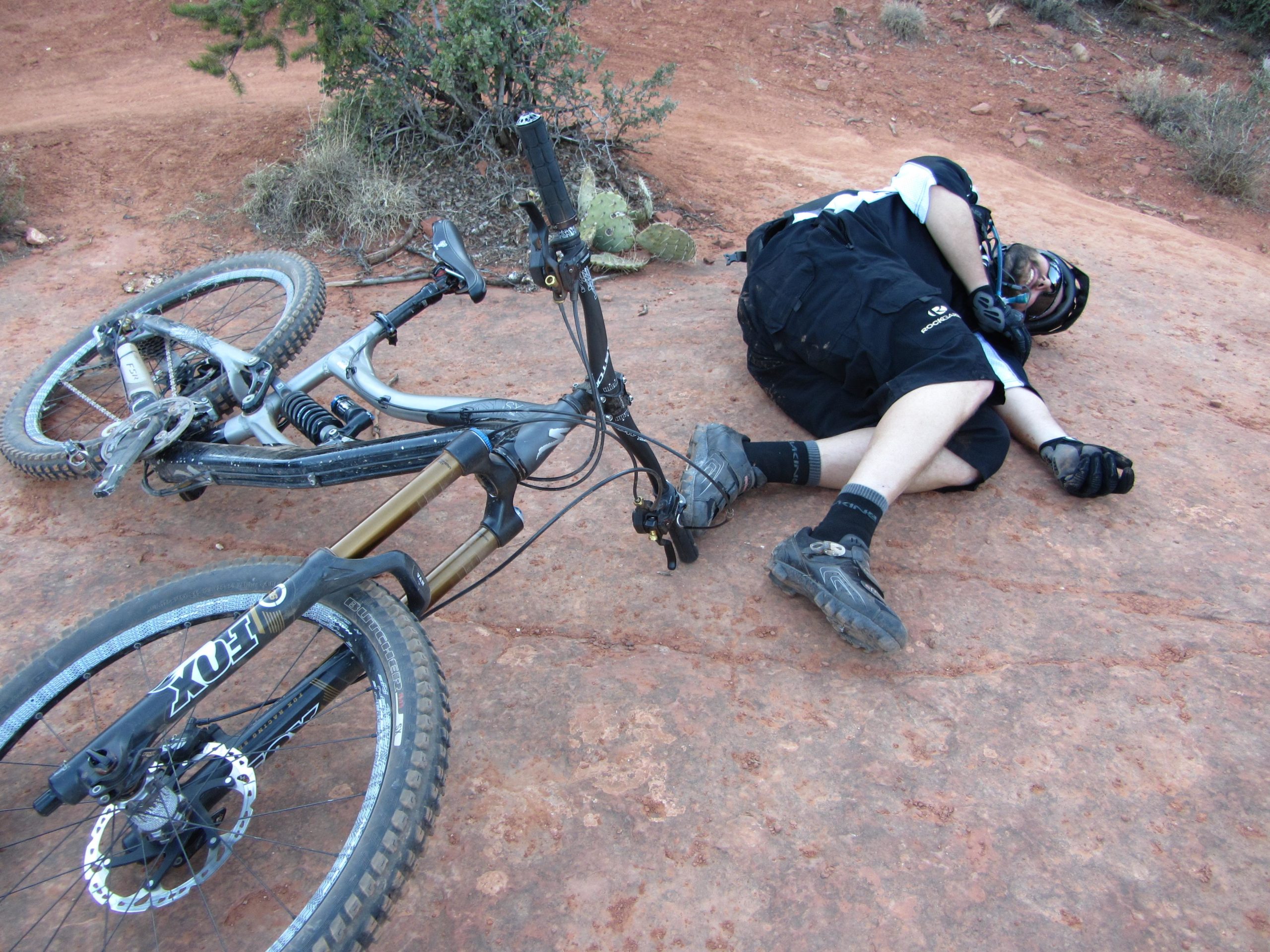 A mountain biker lies on the ground beside an overturned bicycle on a rocky trail. The cyclist, wearing protective gear, appears to be resting or recovering after a fall. The surrounding landscape features red dirt and sparse vegetation typical of a rugged outdoor environment. Bell Rock Area Trails mountain bike trail.