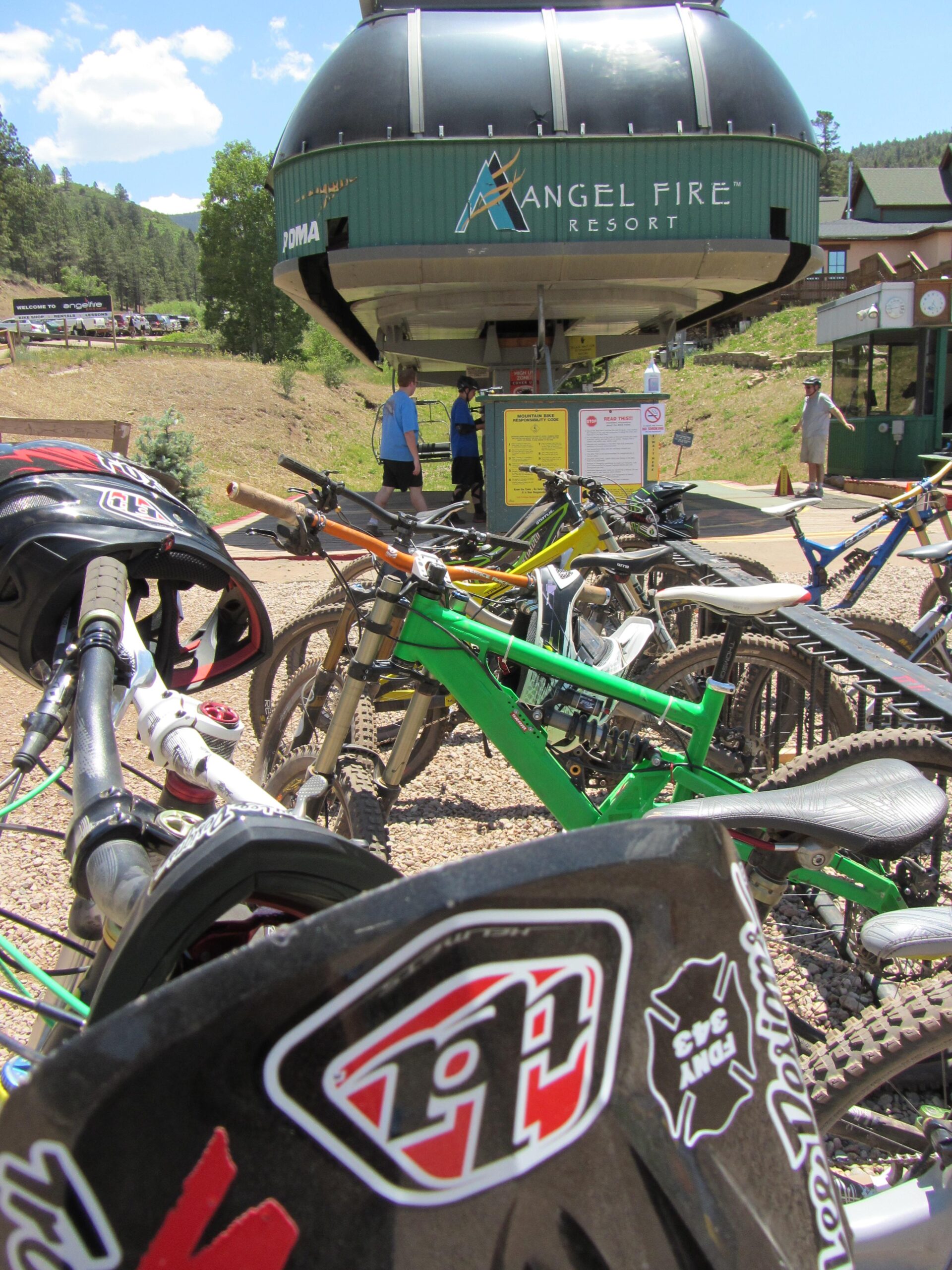 Bicycles parked in front of the Angel Fire Resort's bike lift station on a sunny day, with visitors in the background preparing for a ride. Angel Fire Bike Park mountain bike trail.