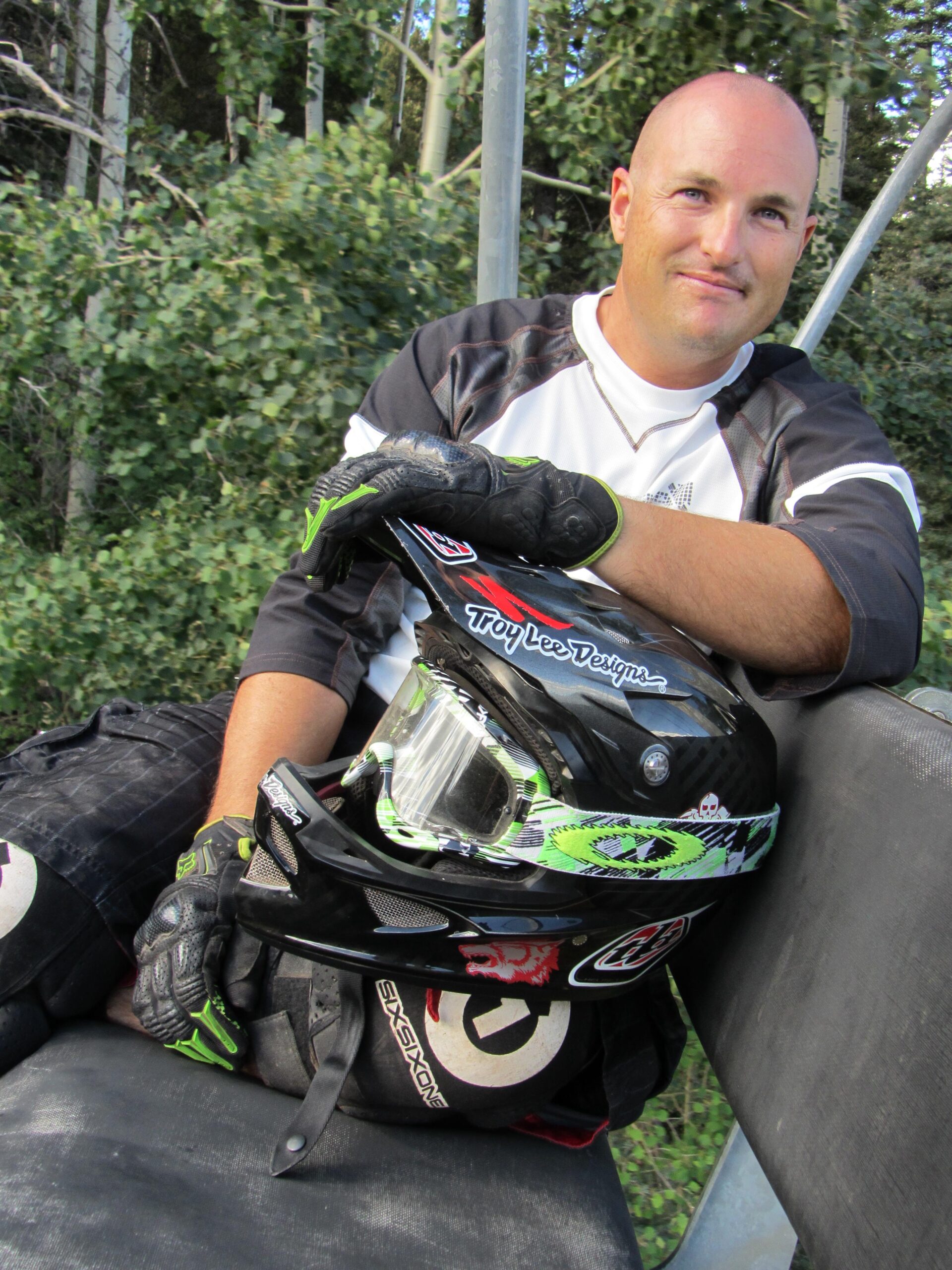 A man sitting on a chairlift wearing a black and white jersey, with protective gloves, leaning casually on a black helmet decorated with green and white designs. He is surrounded by greenery, indicating an outdoor setting, likely in a mountainous or forested area. The atmosphere is relaxed and reflective of a biking or extreme sports environment. Angel Fire Bike Park mountain bike trail.