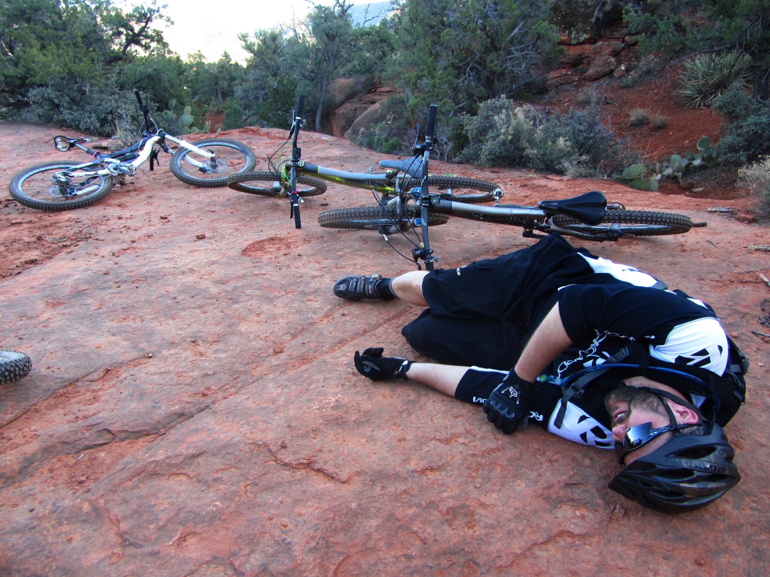 A mountain biker is lying on a rocky surface, appearing to have fallen from their bike, which is positioned nearby on the ground. The scene is set in a rugged outdoor landscape with greenery and rocky terrain in the background. Bell Rock Area Trails mountain bike trail.