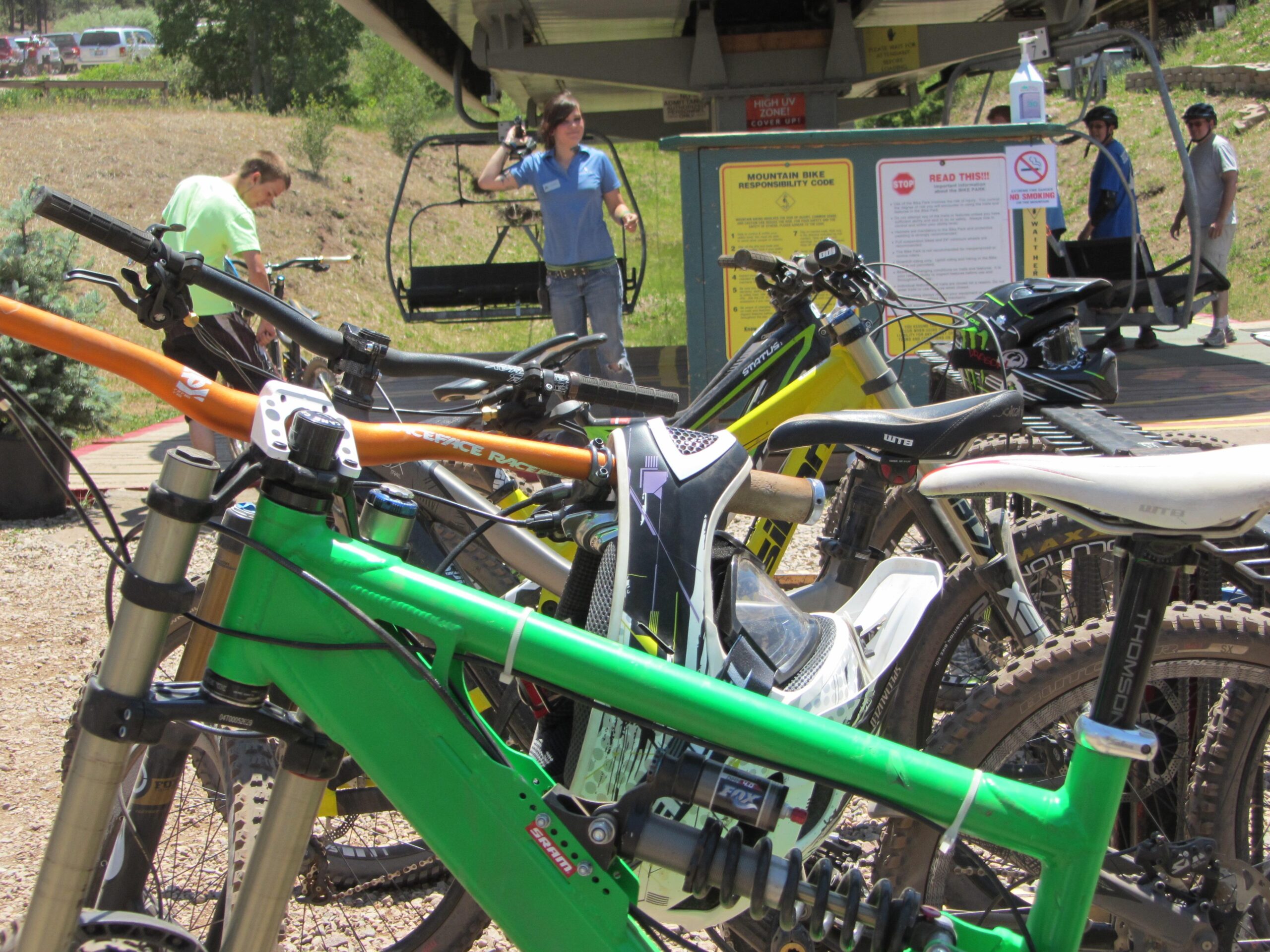 A collection of mountain bikes parked near a ski lift area, with riders and staff in the background. Clear signage about mountain bike safety and codes is visible. The scene is set in a sunny outdoor environment, surrounded by trees and grassy hills. Angel Fire Bike Park mountain bike trail.