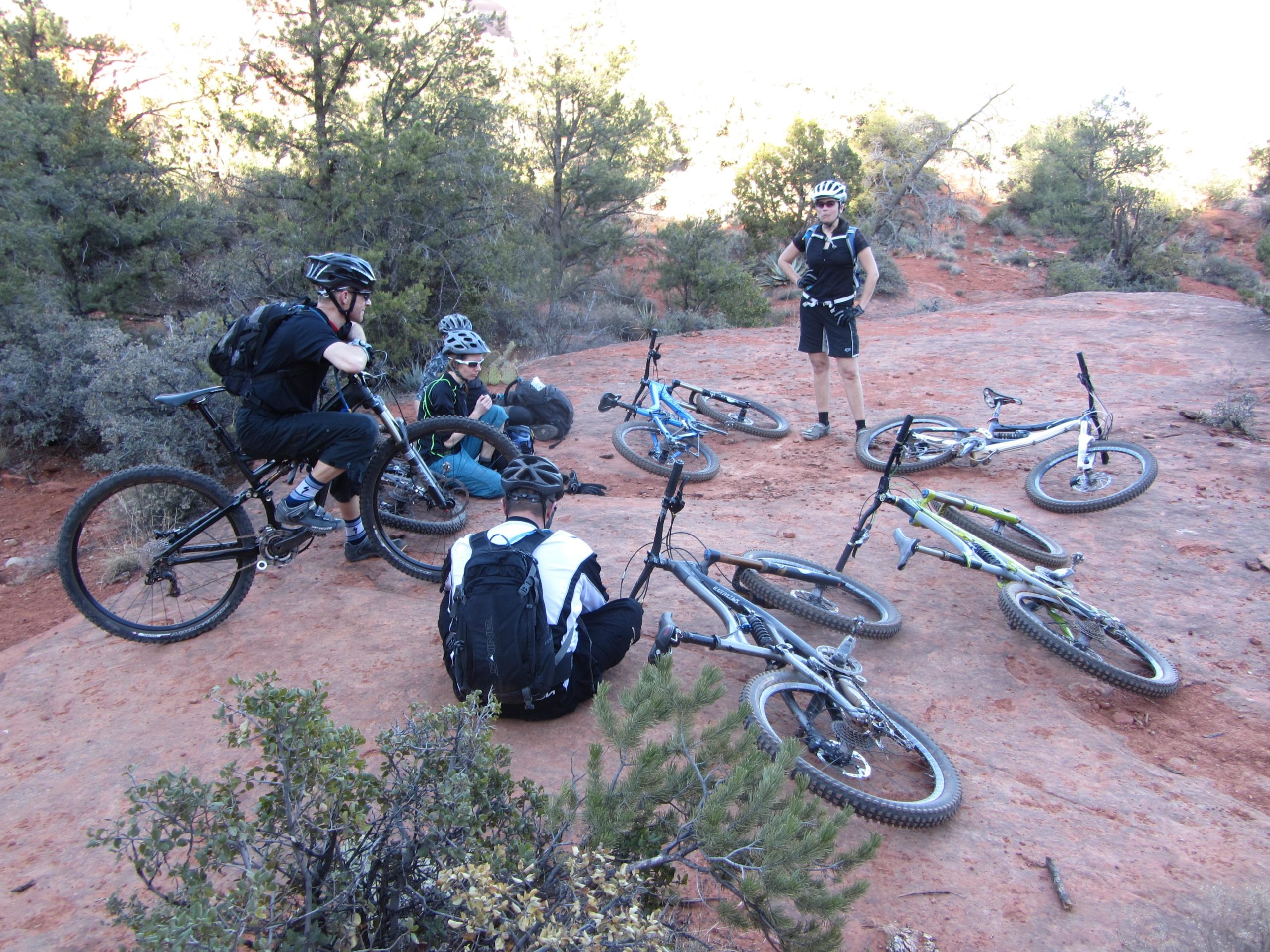 Five mountain bikers take a break on a rocky trail surrounded by shrubs and trees. Several bicycles are laid out around them on the red dirt, while two cyclists are crouched down, inspecting a bike. The group, dressed in cycling gear and helmets, appears to be engaged in discussion, with one member standing upright, hands on hips, observing the others. The scene captures the camaraderie and adventurous spirit of mountain biking. Bell Rock Area Trails mountain bike trail.