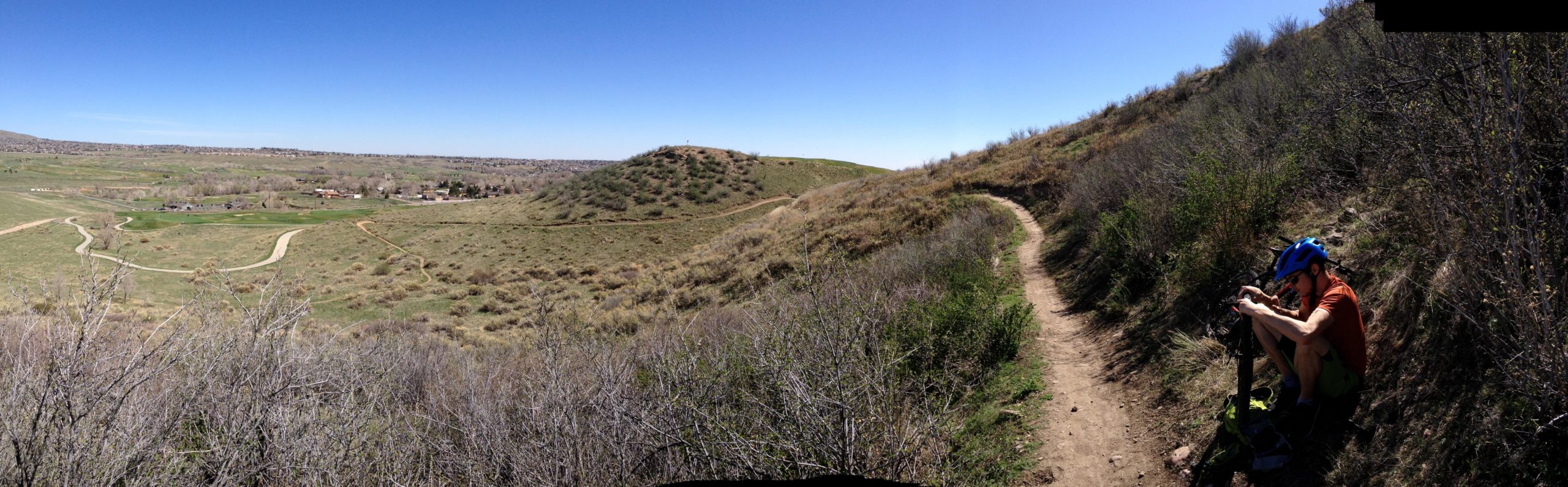 A mountain biker in a blue helmet and orange shirt repairs their bike on a dirt path amidst rolling green hills and sparse vegetation under a clear blue sky. The scenic landscape features winding trails and distant homes in the valley below. Bear Creek Lake Park mountain bike trail.
