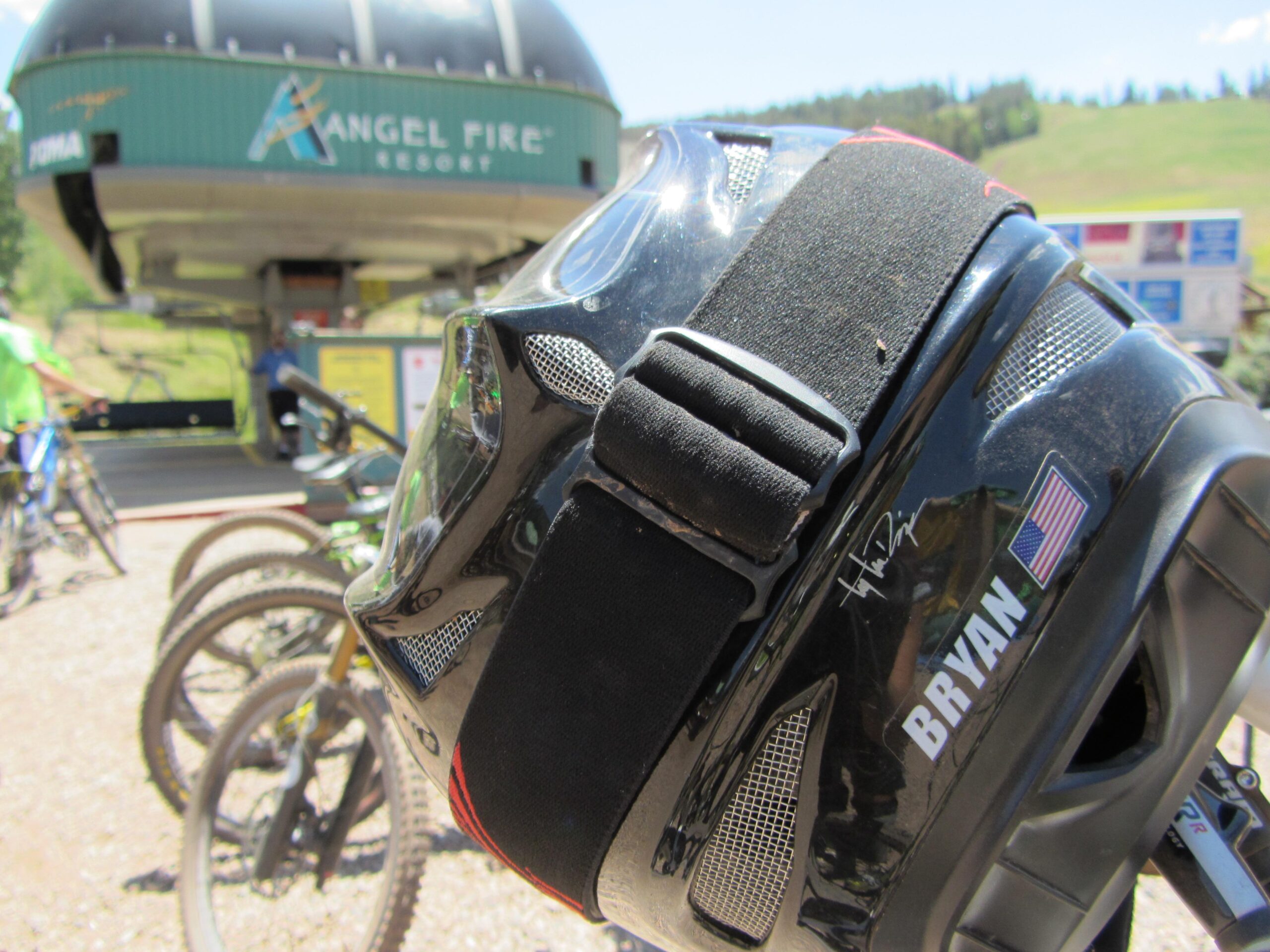 A close-up view of a black bicycle helmet with a name tag and an American flag sticker, positioned in the foreground. In the background, a ski lift station at Angel Fire Resort is visible, with people and mountain bikes nearby. The scene is set in a sunny outdoor environment with green hills in the distance. Angel Fire Bike Park mountain bike trail.