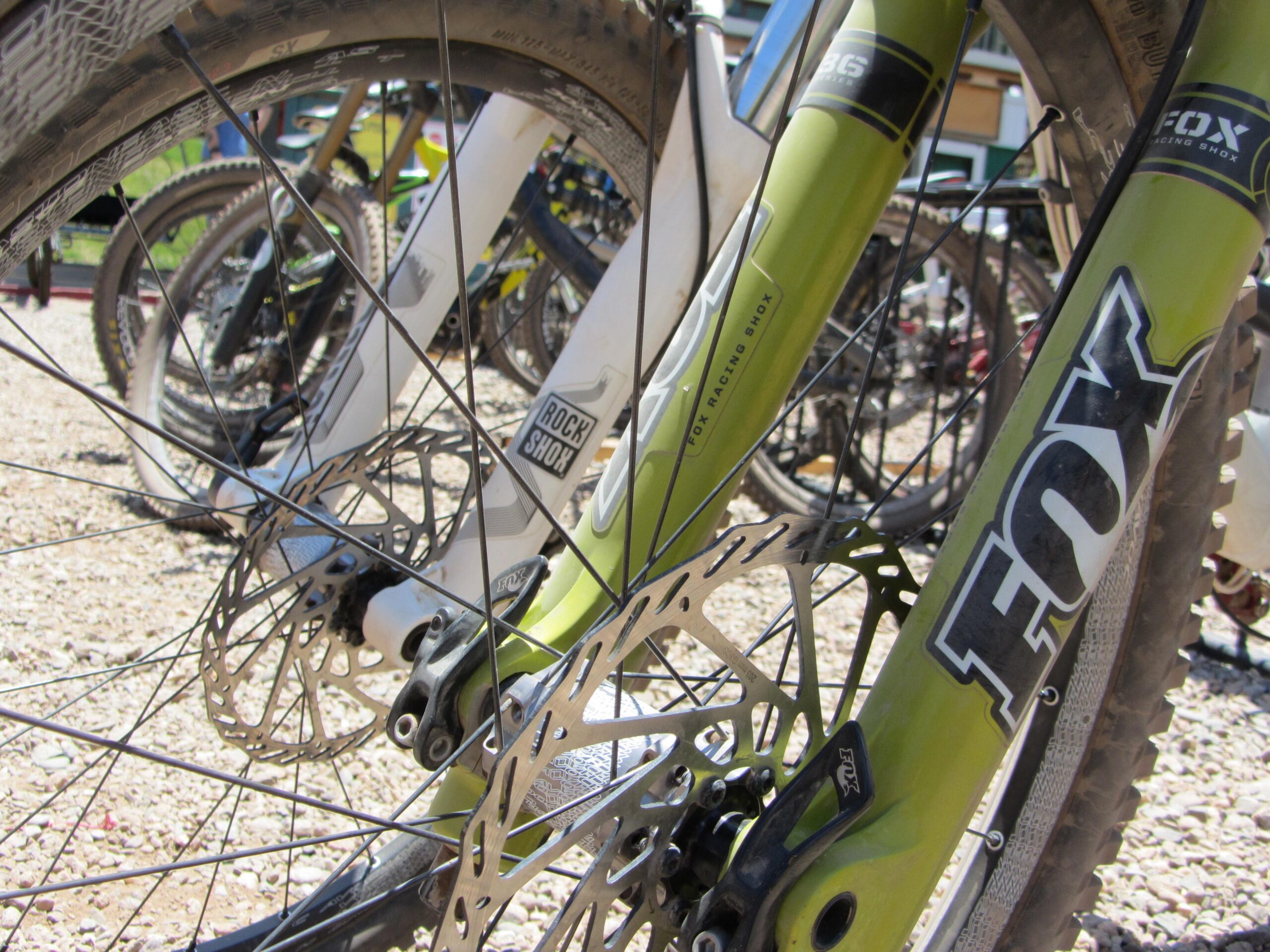 Close-up of the front fork and wheel assembly of a mountain bike, featuring a green FOX suspension fork and disc brake setup. The background shows additional bicycles parked on a gravel surface under bright sunlight. Angel Fire Bike Park mountain bike trail.