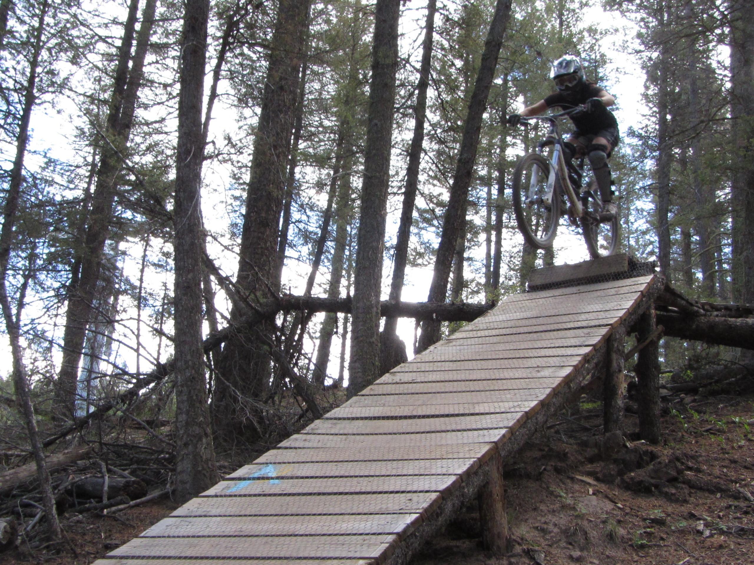 A mountain biker in protective gear performs a jump off a wooden ramp in a forested area, surrounded by tall trees and natural foliage. The scene captures the action of the biker mid-air as they navigate a challenging trail. Angel Fire Bike Park mountain bike trail.