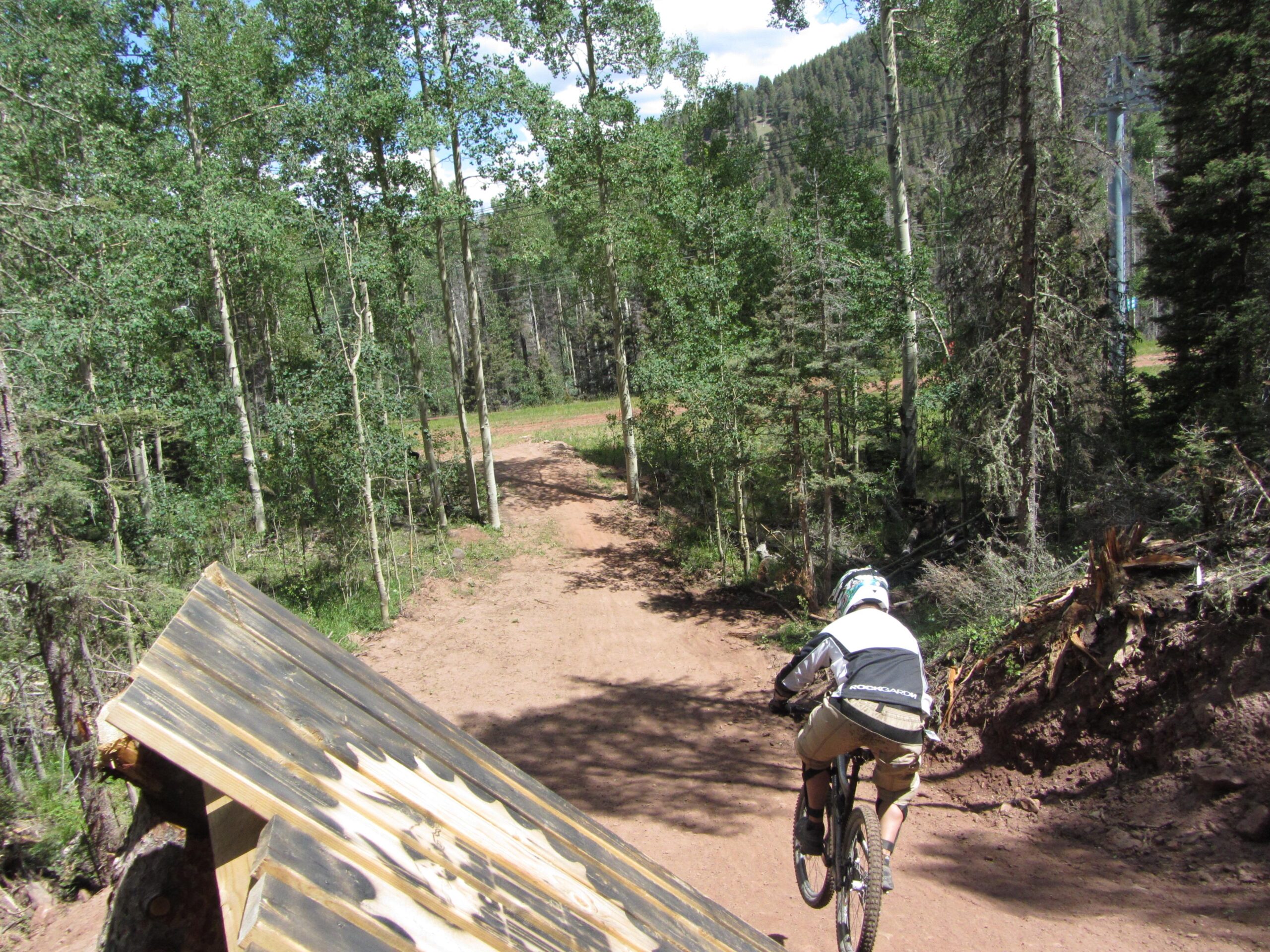 A mountain biker descends from a wooden ramp onto a dirt path surrounded by tall trees in a forested area, with a clear sky visible in the background. Angel Fire Bike Park mountain bike trail.