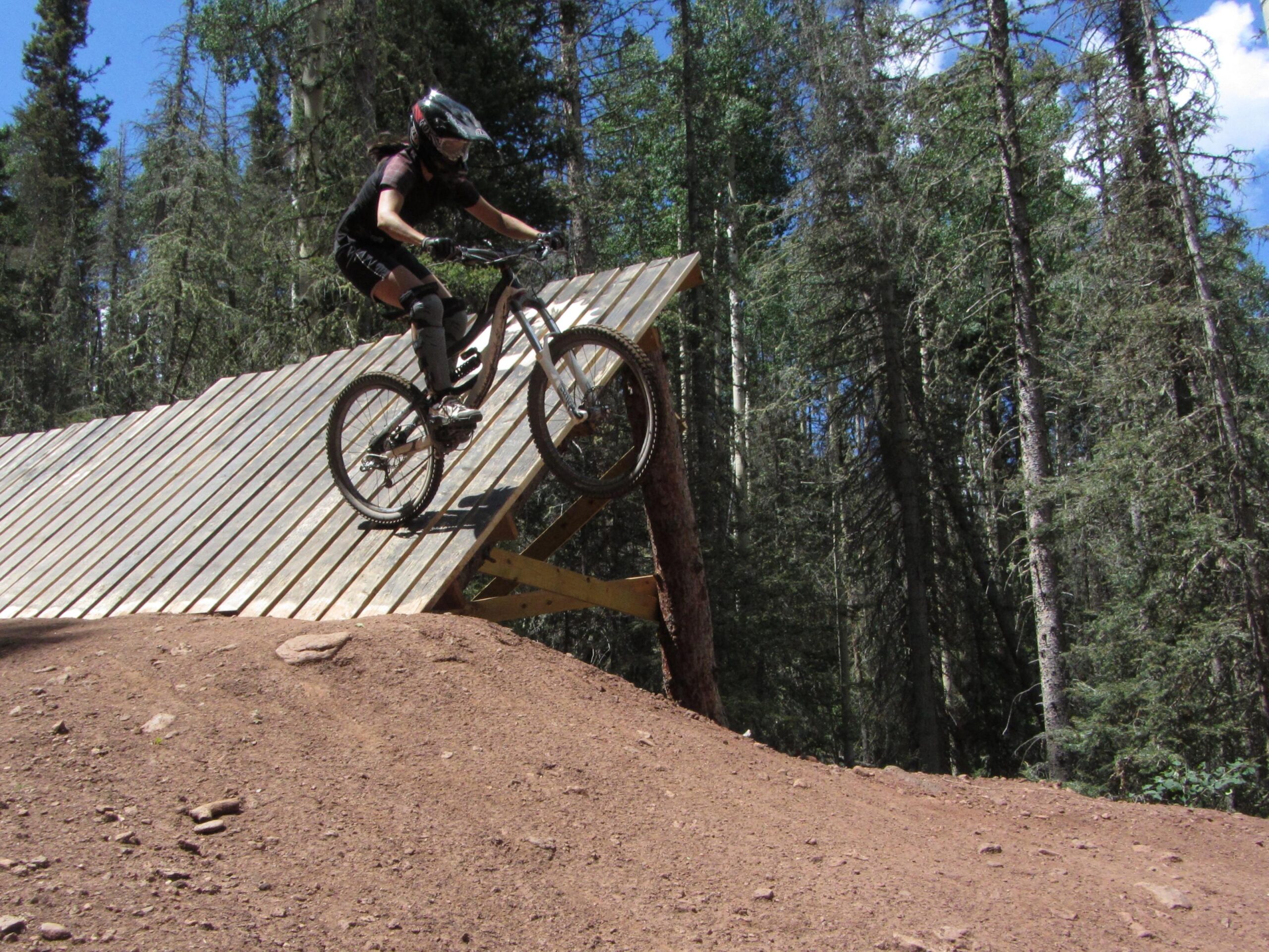 A mountain biker in protective gear is mid-air as they descend from a wooden ramp in a forested area. The bike is in motion, showcasing the rider's skill, with trees in the background and a clear blue sky above. Angel Fire Bike Park mountain bike trail.