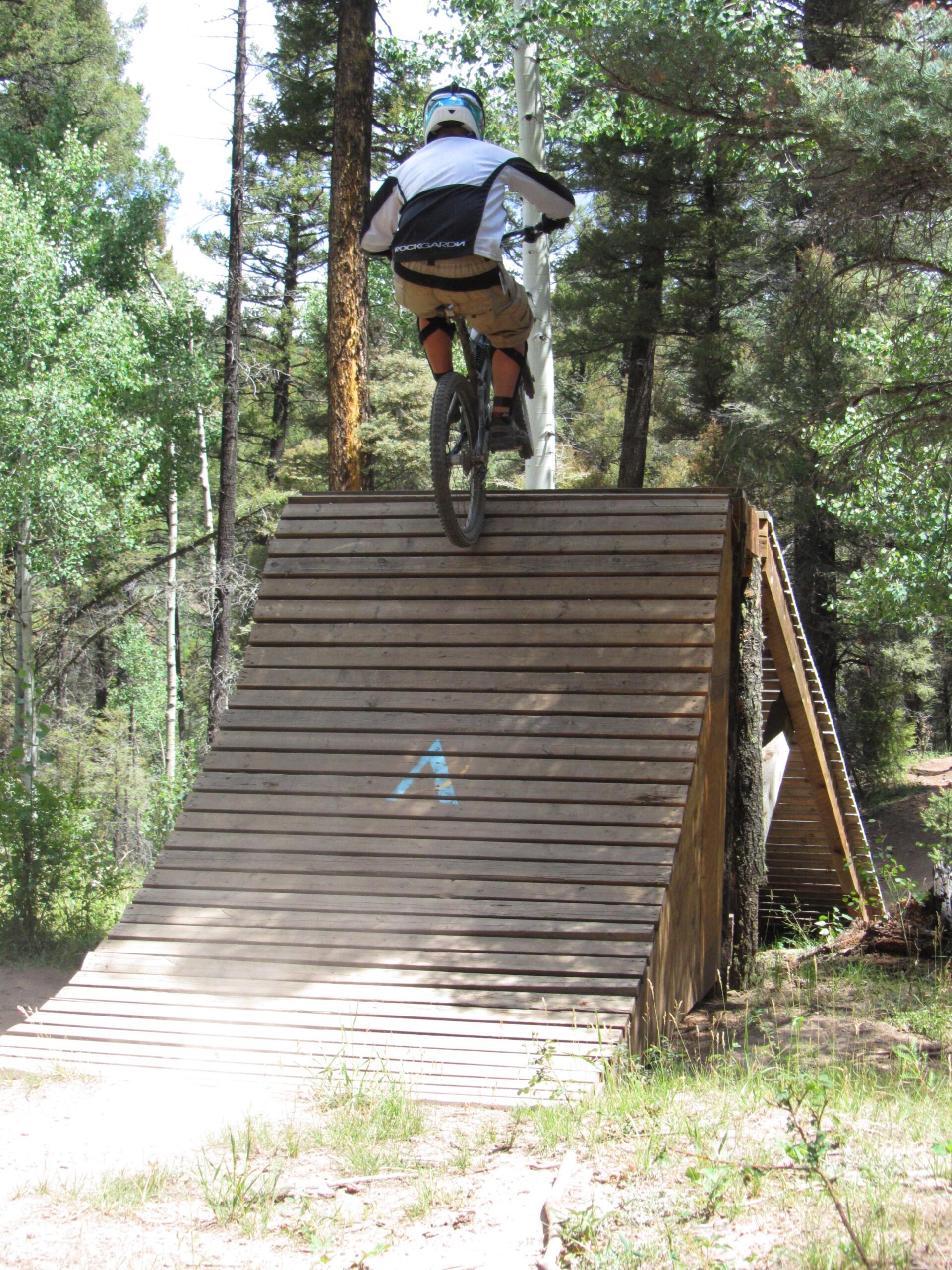 A mountain biker in a helmet and protective gear is seen descending a wooden ramp in a forested area, with trees and greenery in the background. Dust rises from the ground as the biker approaches the bottom of the ramp. Angel Fire Bike Park mountain bike trail.
