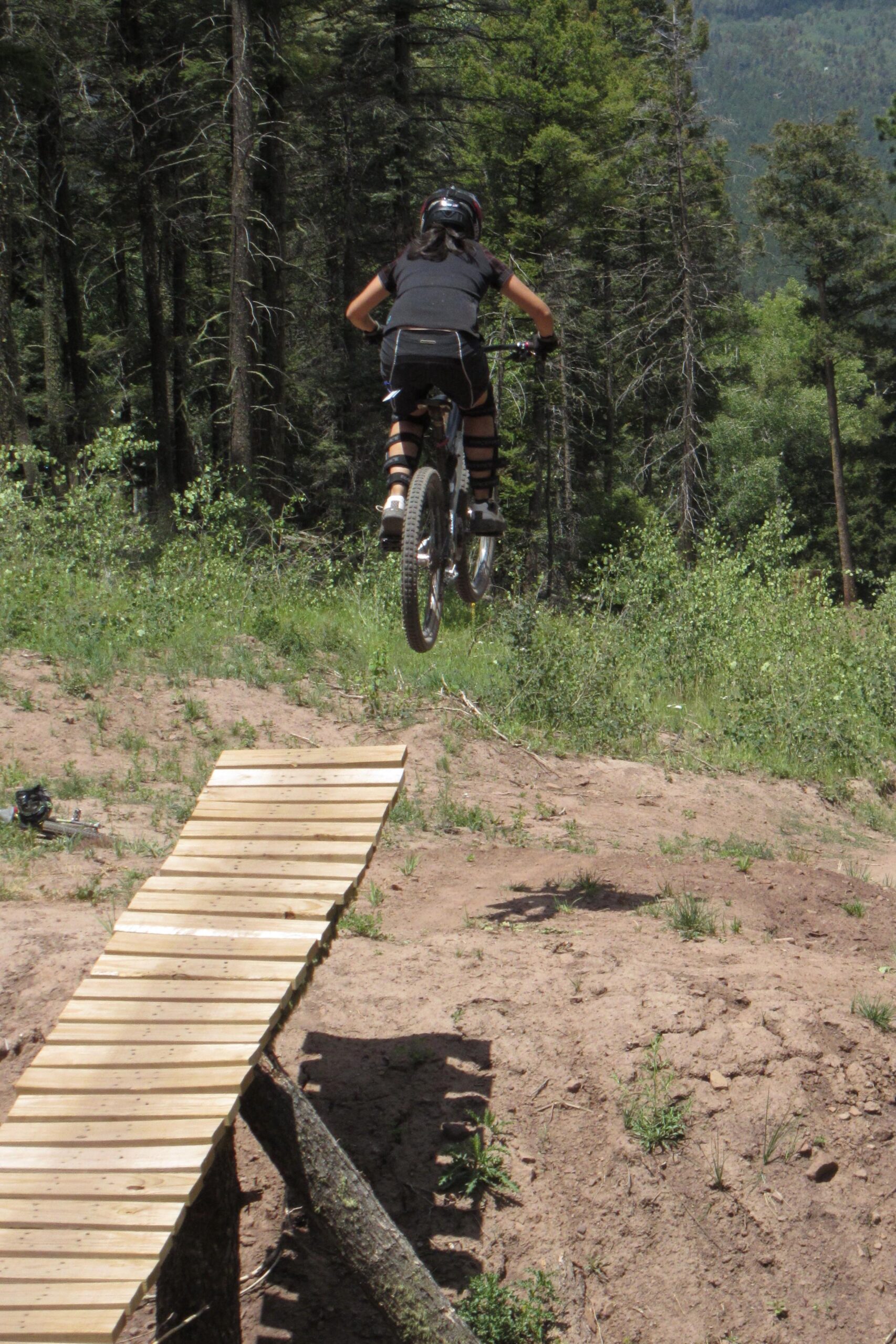 A person riding a mountain bike jumps off a wooden ramp in a forested area, with trees in the background and a grassy terrain. Angel Fire Bike Park mountain bike trail.
