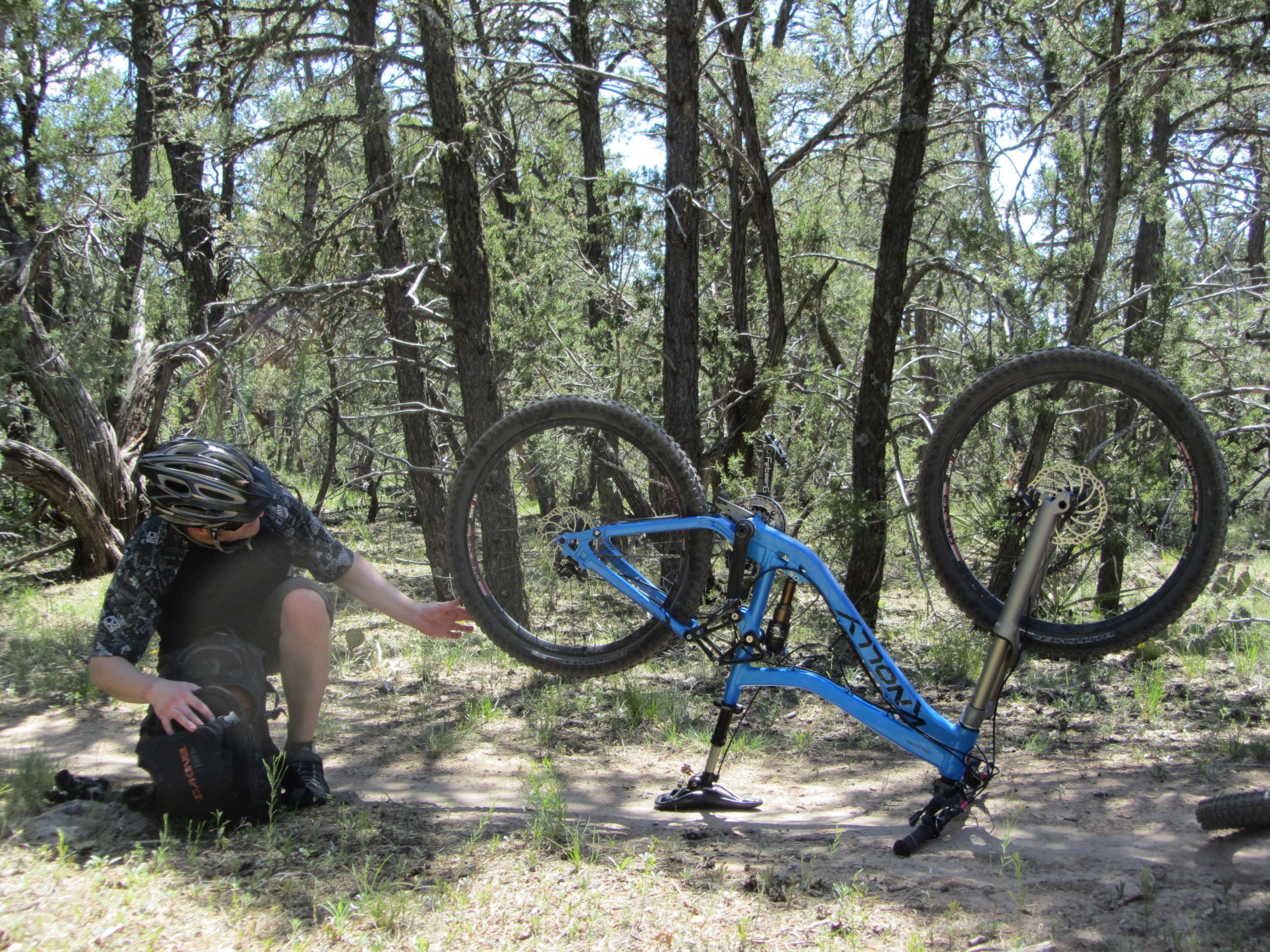 A mountain biker in a helmet and casual attire is crouching beside a black backpack in a forested area. Nearby, a blue mountain bike is upside down, propped up on its handlebars and rear tire, with trees and greenery in the background. Otero Canyon mountain bike trail.