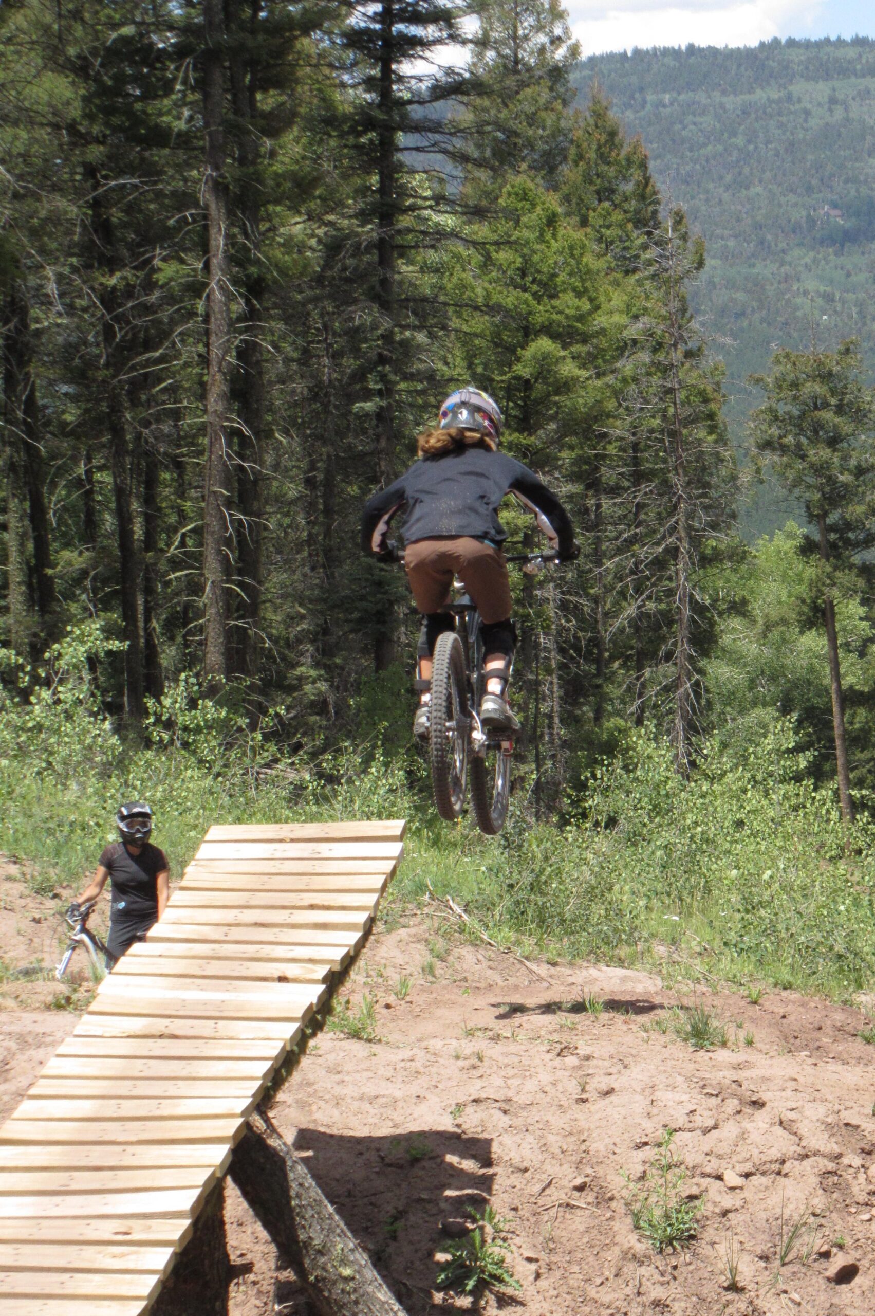 A person in a black shirt and brown shorts jumps off a wooden bike ramp while riding a mountain bike, with trees and mountains in the background. Another individual in a black helmet and shirt stands nearby, watching the jump. Angel Fire Bike Park mountain bike trail.