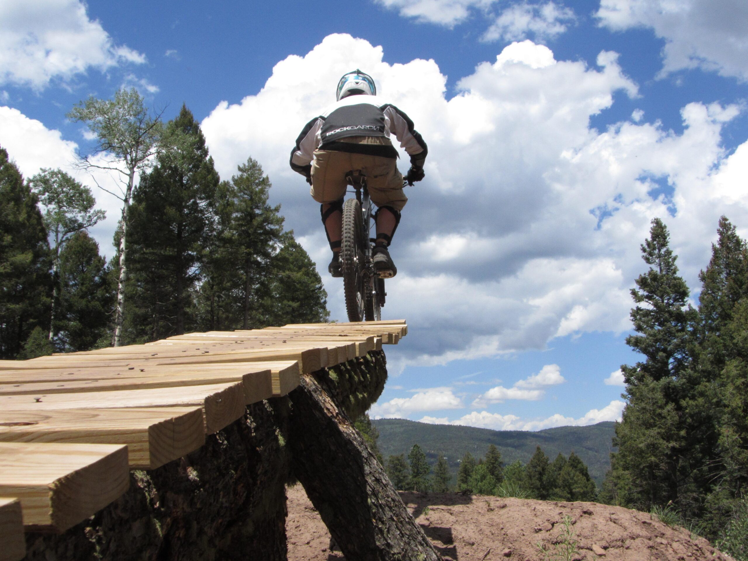 A cyclist riding a mountain bike on a wooden plank bridge in a forested area, with trees in the background and a blue sky filled with clouds. The view captures the thrill of outdoor biking and the natural landscape. Angel Fire Bike Park mountain bike trail.