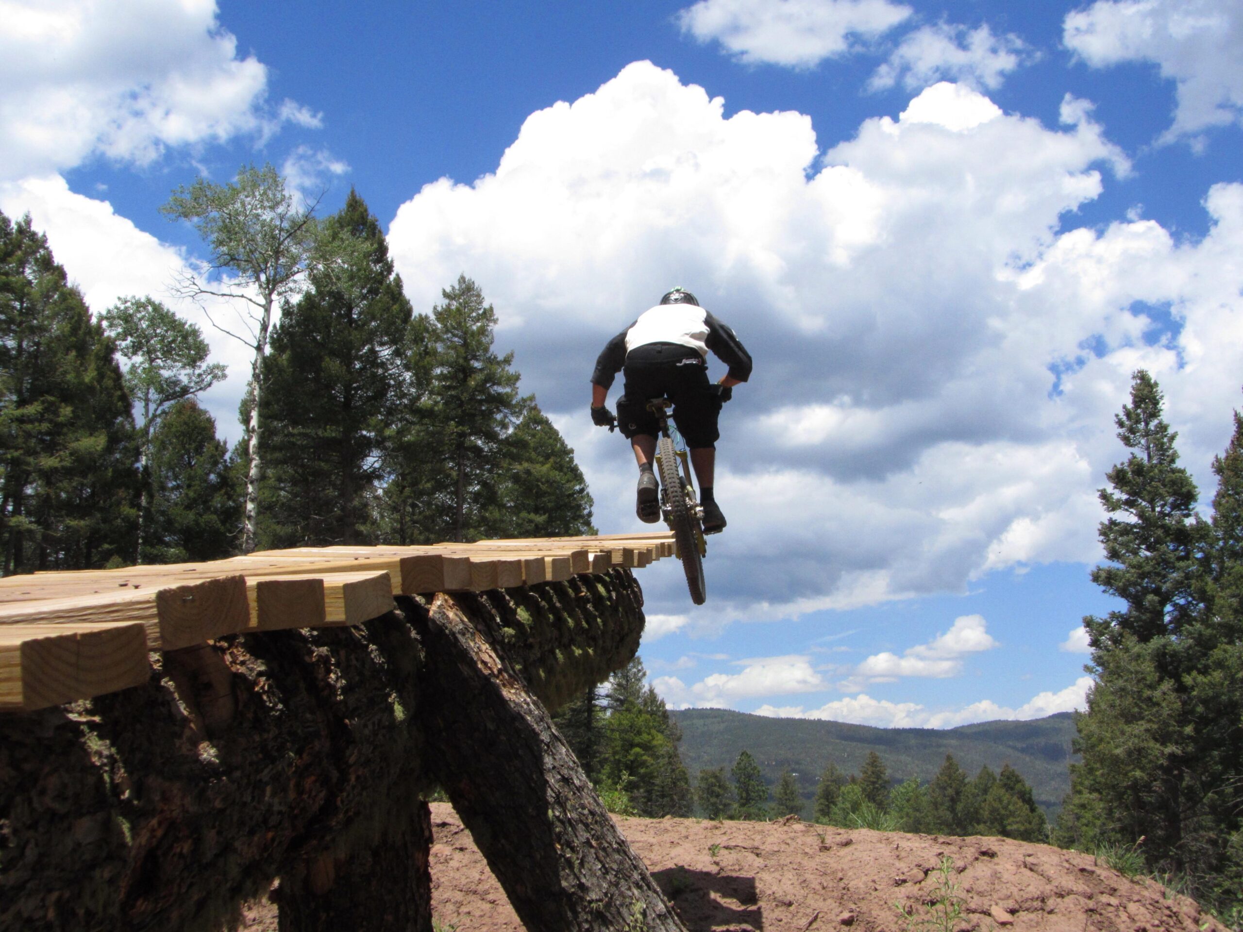 A mountain biker catches air while jumping off a wooden ramp suspended over a scenic landscape, surrounded by tall trees and a blue sky filled with clouds. Angel Fire Bike Park mountain bike trail.