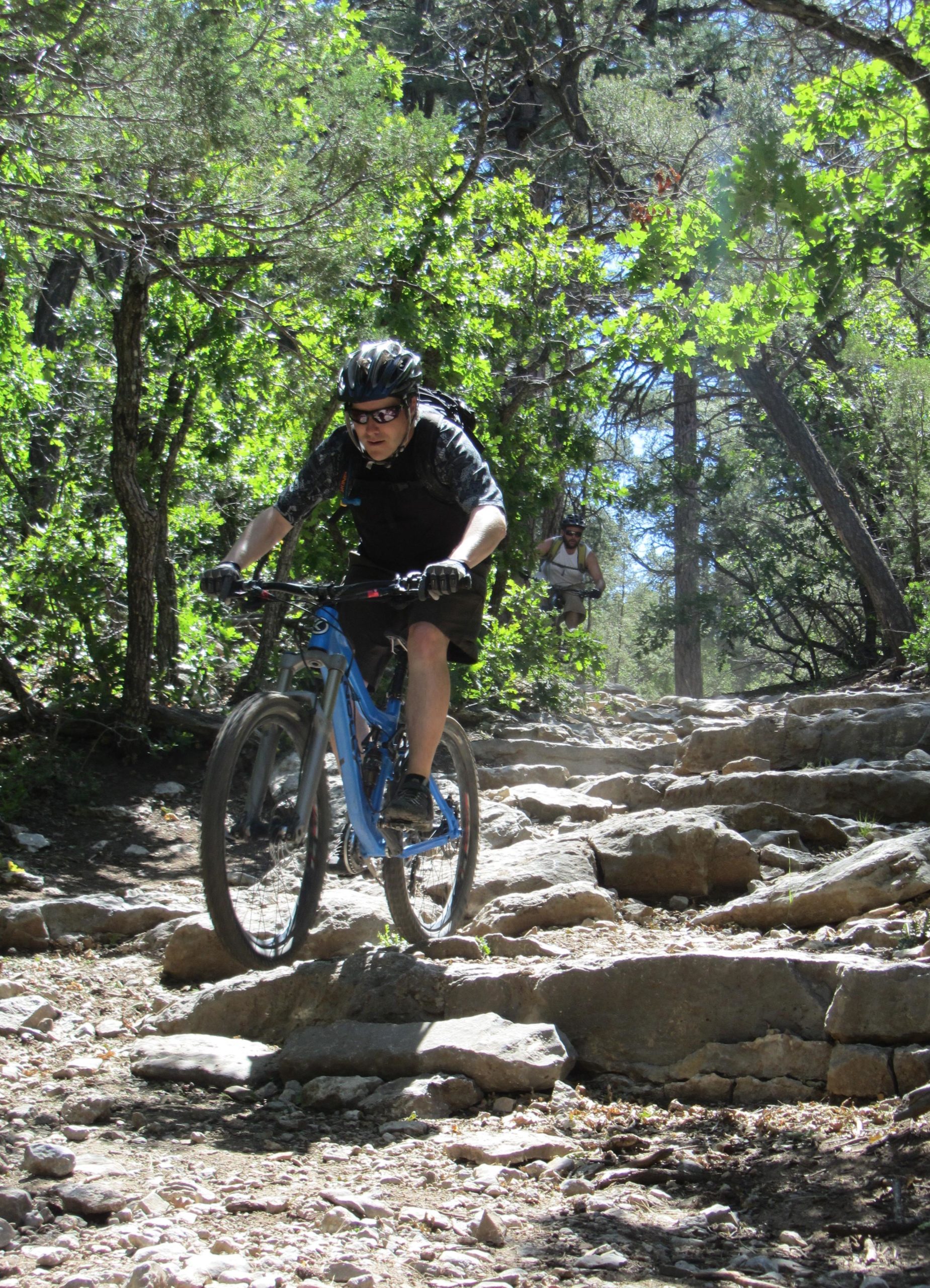A mountain biker navigating a rocky trail in a wooded area, wearing a helmet and sunglasses. In the background, another cyclist can be seen further up the trail. The sunlight filters through the trees, highlighting the rugged terrain. Otero Canyon mountain bike trail.
