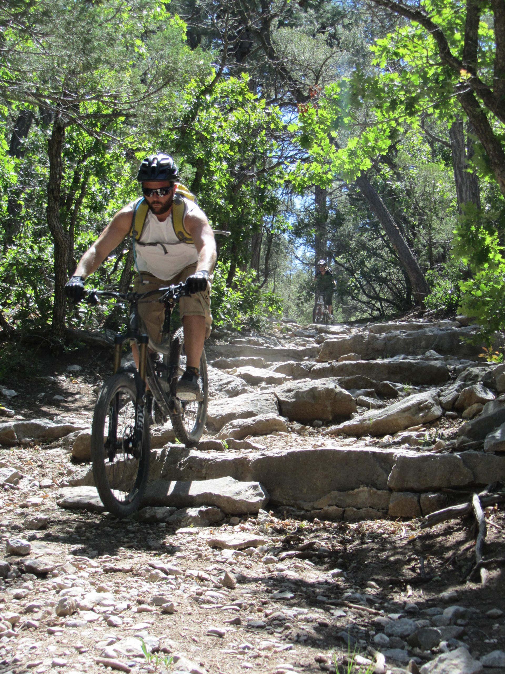 A mountain biker riding on a rocky trail surrounded by lush green trees, with another cyclist visible in the background. The sun is shining, creating a bright and vibrant outdoor atmosphere. Otero Canyon mountain bike trail.