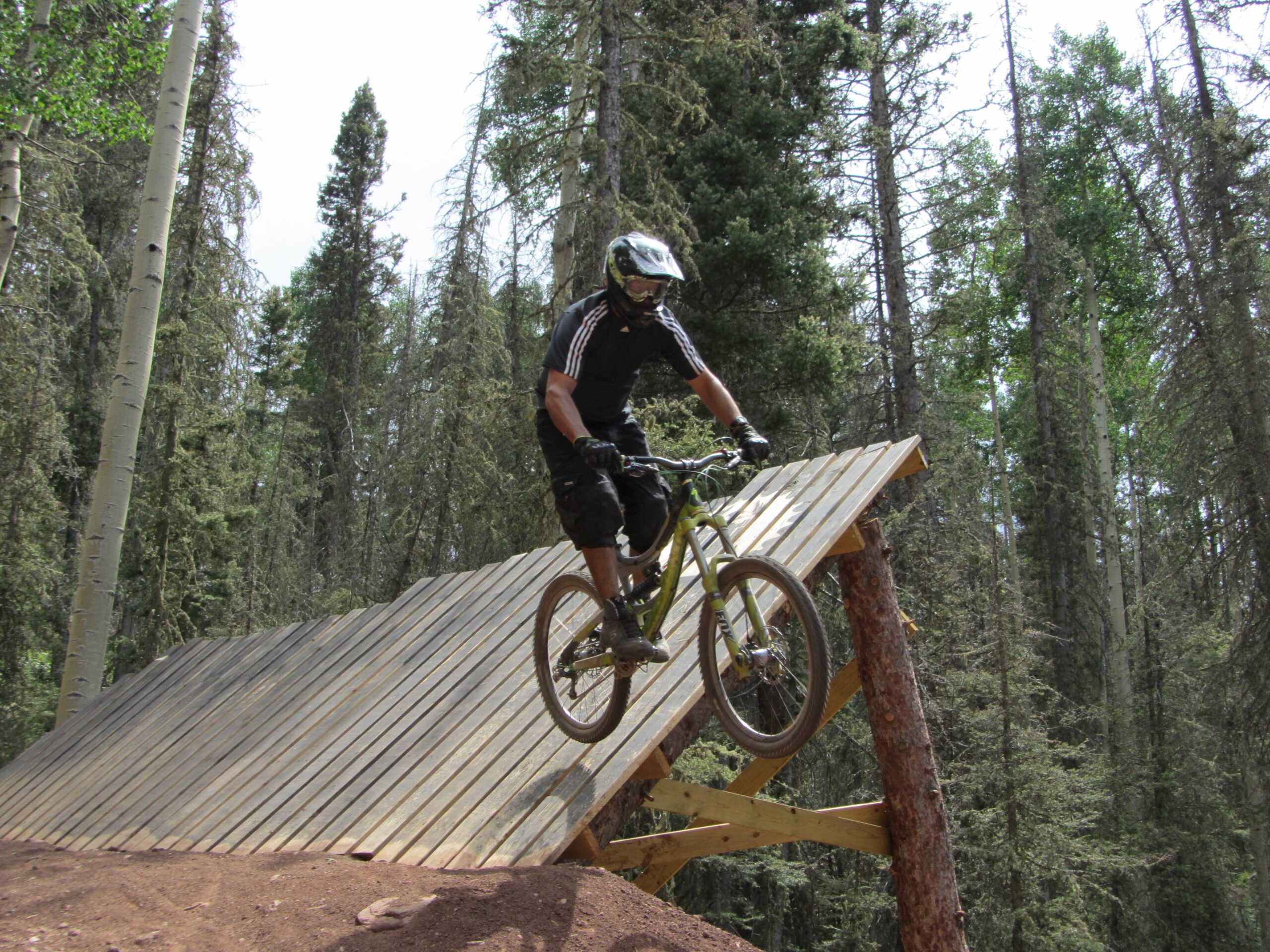 A mountain biker wearing a helmet and protective gear rides down a wooden ramp in a forested area. The bike is airborne as the rider approaches the edge of the ramp, surrounded by tall trees and natural scenery. Angel Fire Bike Park mountain bike trail.