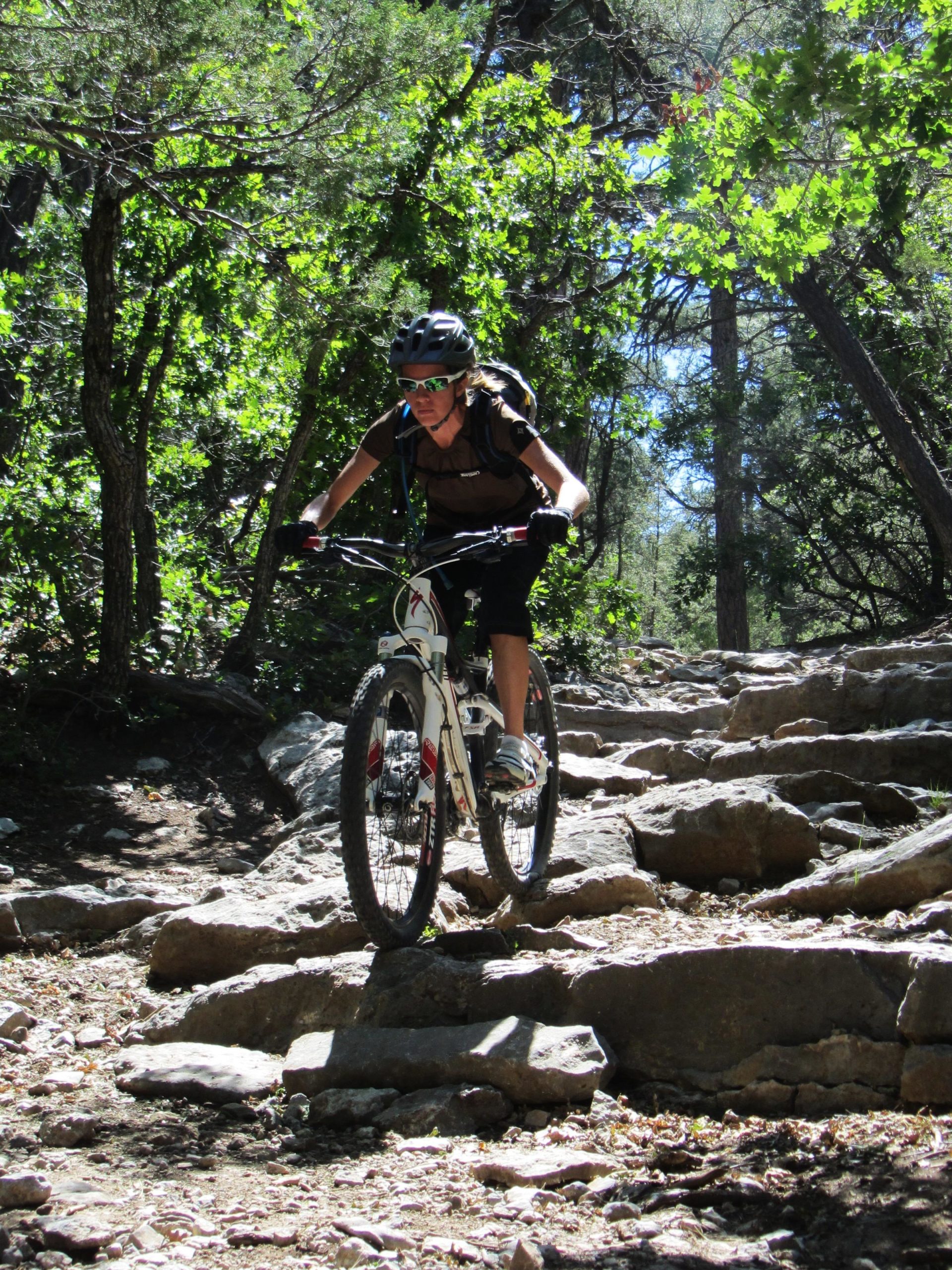 A mountain biker navigating a rocky trail surrounded by trees, wearing a helmet and sunglasses. The cyclist is focused as they tackle the uneven terrain, showcasing a dynamic moment of outdoor adventure. Otero Canyon mountain bike trail.