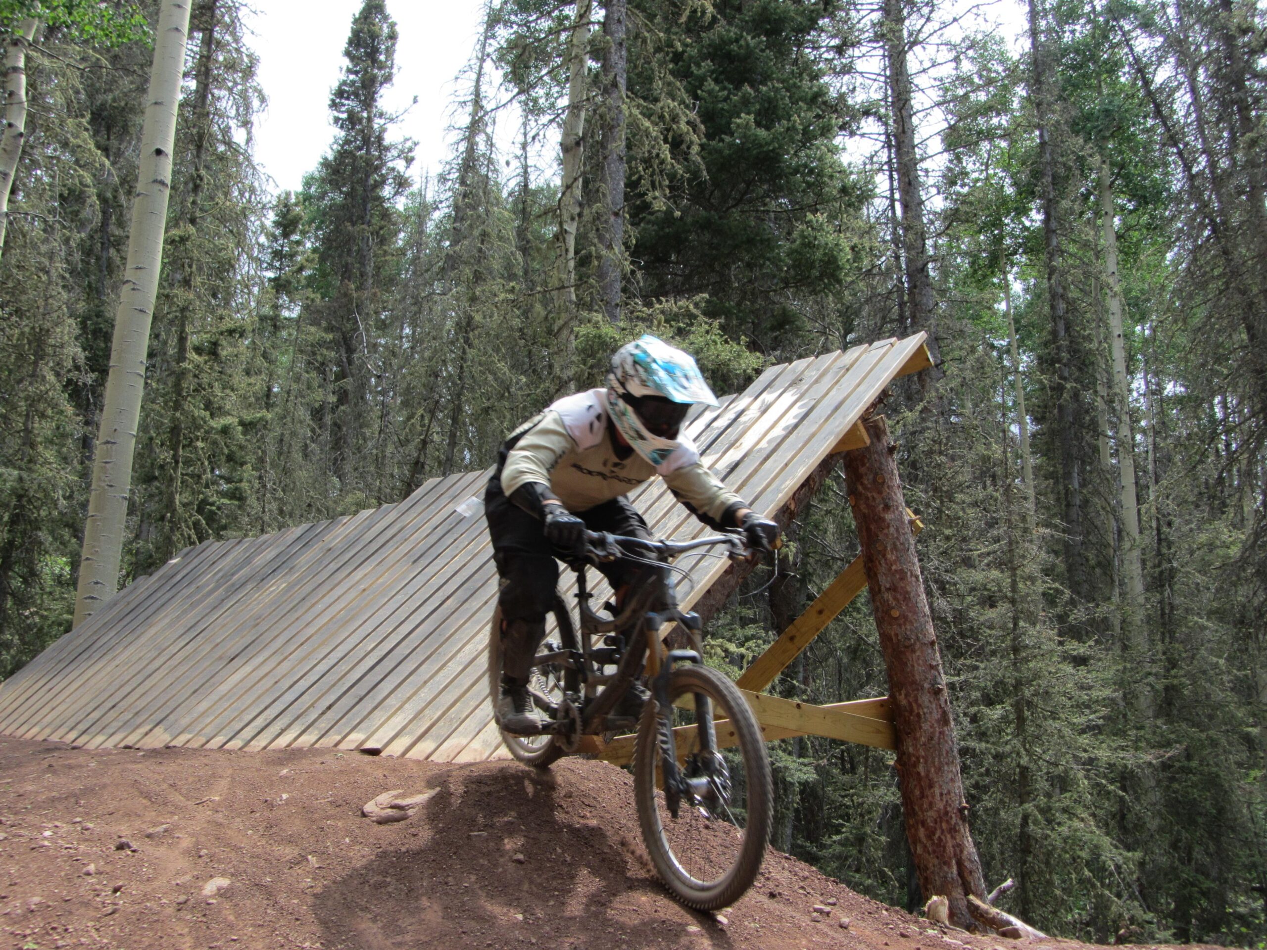 A mountain biker in protective gear is descending a wooden ramp in a wooded area, surrounded by tall trees. The rider leans into the turn while navigating a dirt trail. The scene captures the thrill of off-road biking in a natural setting. Angel Fire Bike Park mountain bike trail.