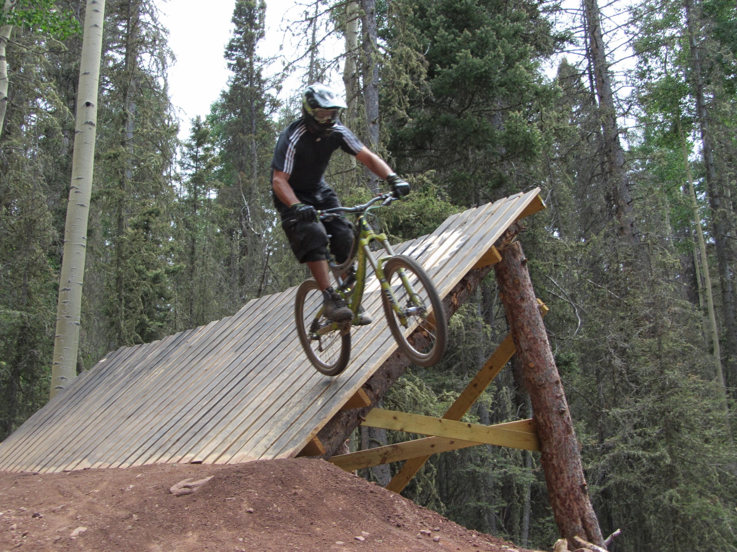 A mountain biker in mid-air performing a jump off a wooden ramp, surrounded by tall trees in a forested area. The biker is wearing a helmet and protective gear, showcasing an energetic and dynamic action shot. Angel Fire Bike Park mountain bike trail.