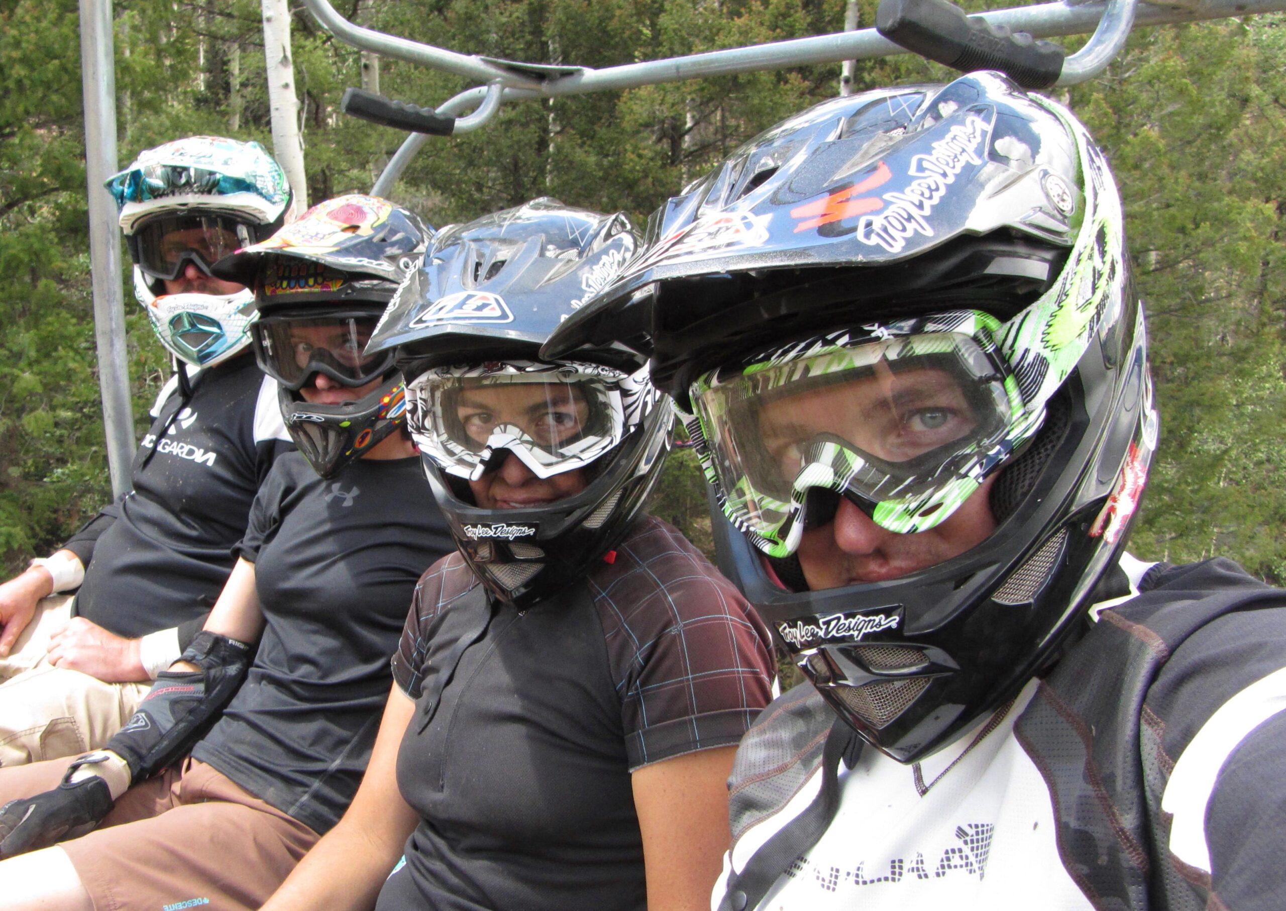 A group of four individuals wearing protective helmets and goggles sitting on a ski lift chair, surrounded by green trees. They are dressed in athletic gear suitable for biking or skiing. The image captures their focus and anticipation as they prepare for an outdoor adventure. Angel Fire Bike Park mountain bike trail.