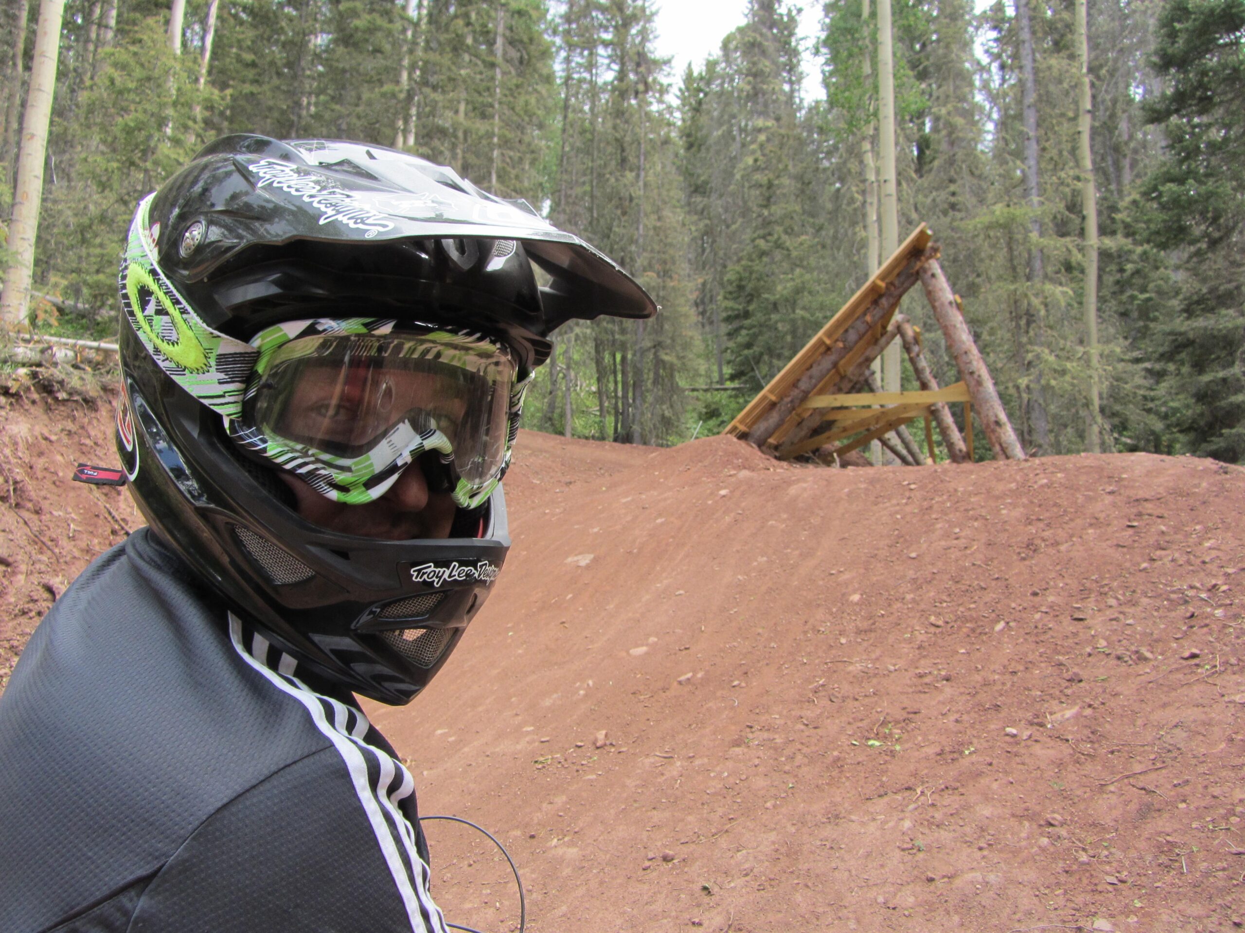 A person wearing a black motorcycle helmet and goggles sits near a dirt track in a forested area, with a wooden jump ramp visible in the background. The landscape features tall trees and a dirt surface. Angel Fire Bike Park mountain bike trail.