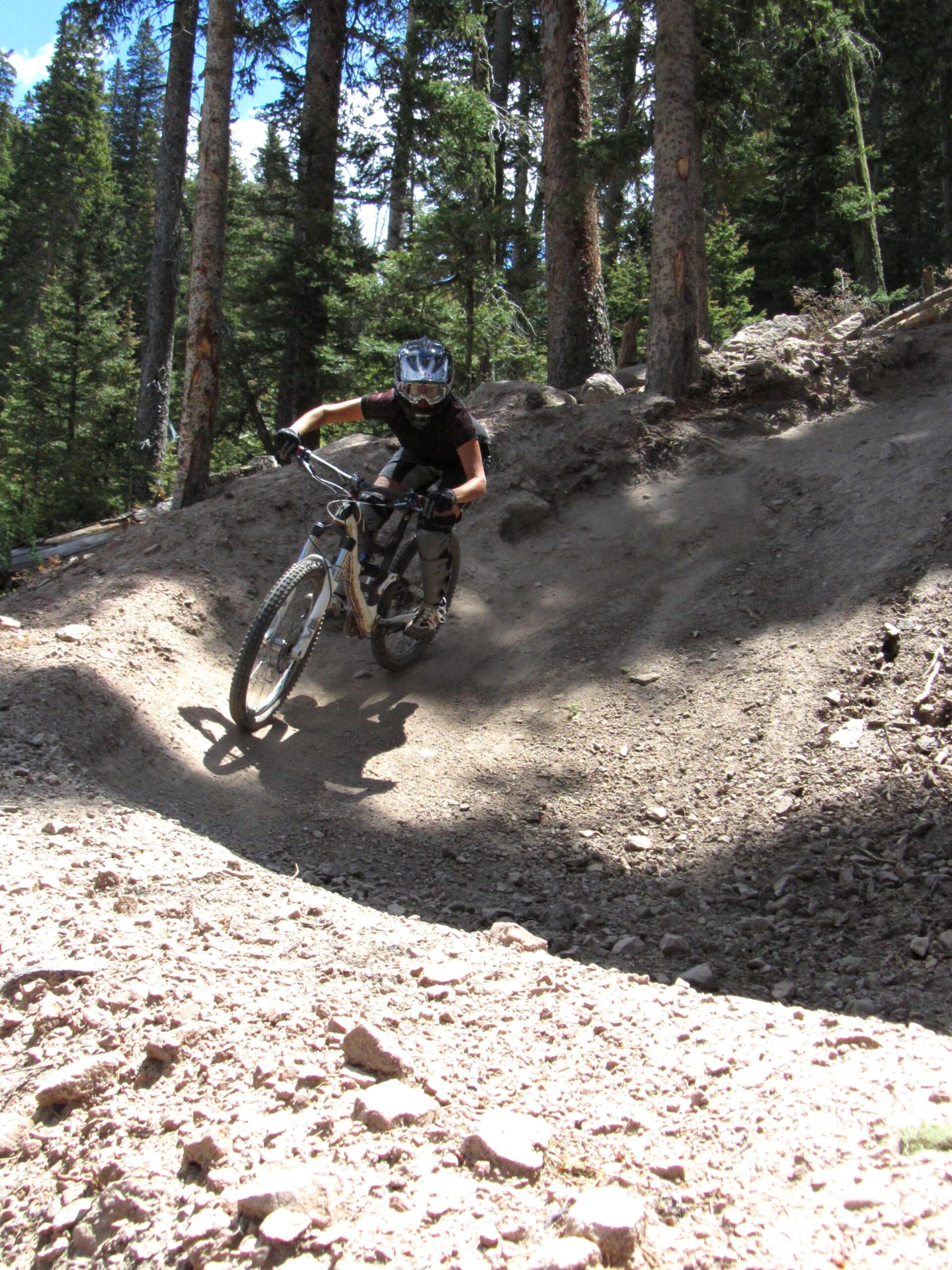 A mountain biker navigating a dirt trail in a forested area, leaning into a turn and kicking up dust. The cyclist is wearing a helmet and protective gear, surrounded by tall trees and blue skies in the background. Angel Fire Bike Park mountain bike trail.