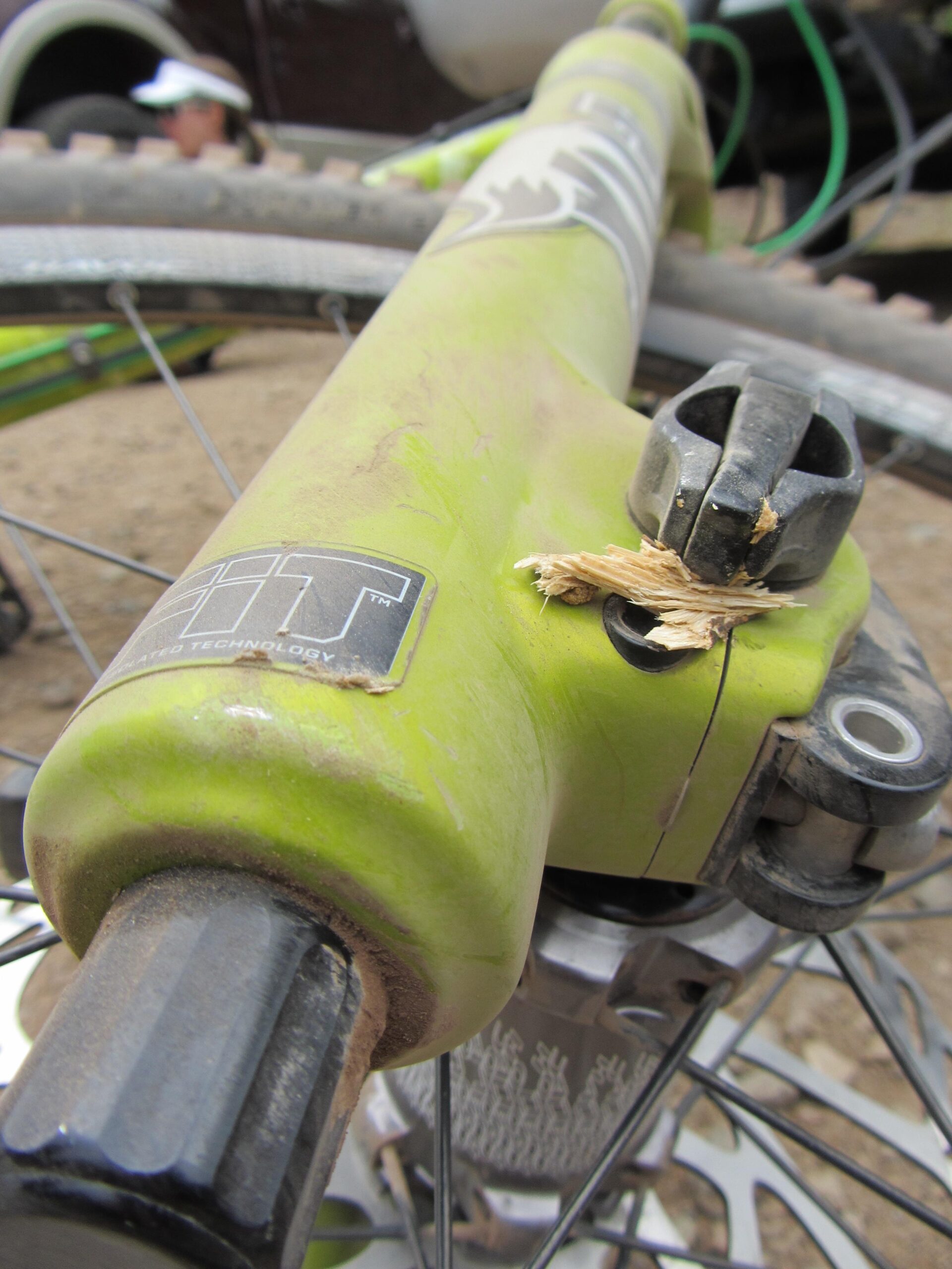 A close-up view of a green bicycle fork, showcasing dirt and scratches, with a small piece of wood lodged in the mechanism. The bike's wheel and spokes are partially visible in the foreground, and a blurred figure in the background suggests an outdoor setting. Angel Fire Bike Park mountain bike trail.