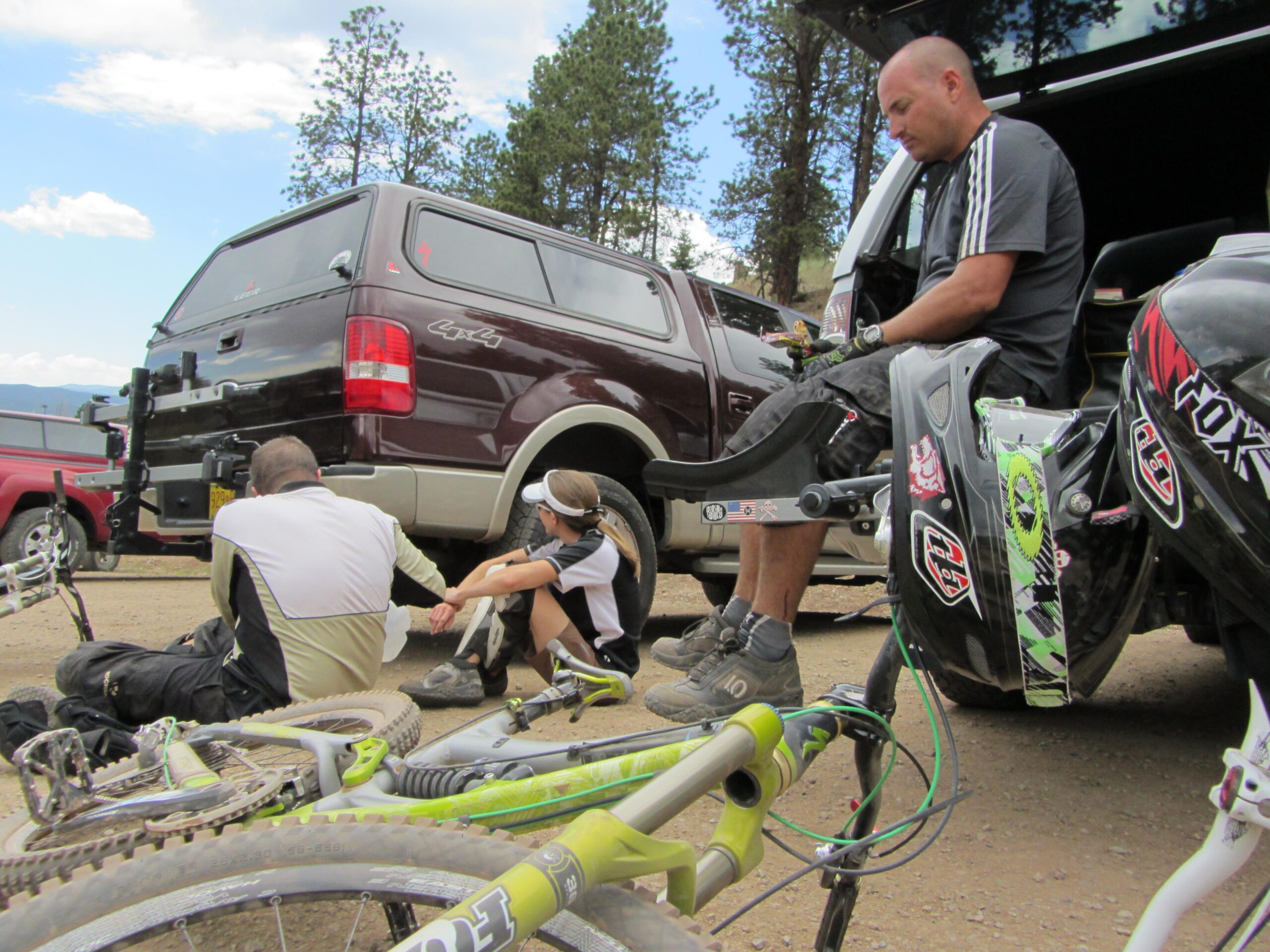 Three individuals are gathered near the back of a parked truck in an outdoor setting, surrounded by trees. Two of them are sitting on the ground, engaged in conversation or relaxation, while the third sits on a bike seat. Mountain bikes are leaning on the ground nearby, showcasing a vibrant green and more muted colors. The sky is partly cloudy, suggesting a nice day for outdoor activities. Angel Fire Bike Park mountain bike trail.