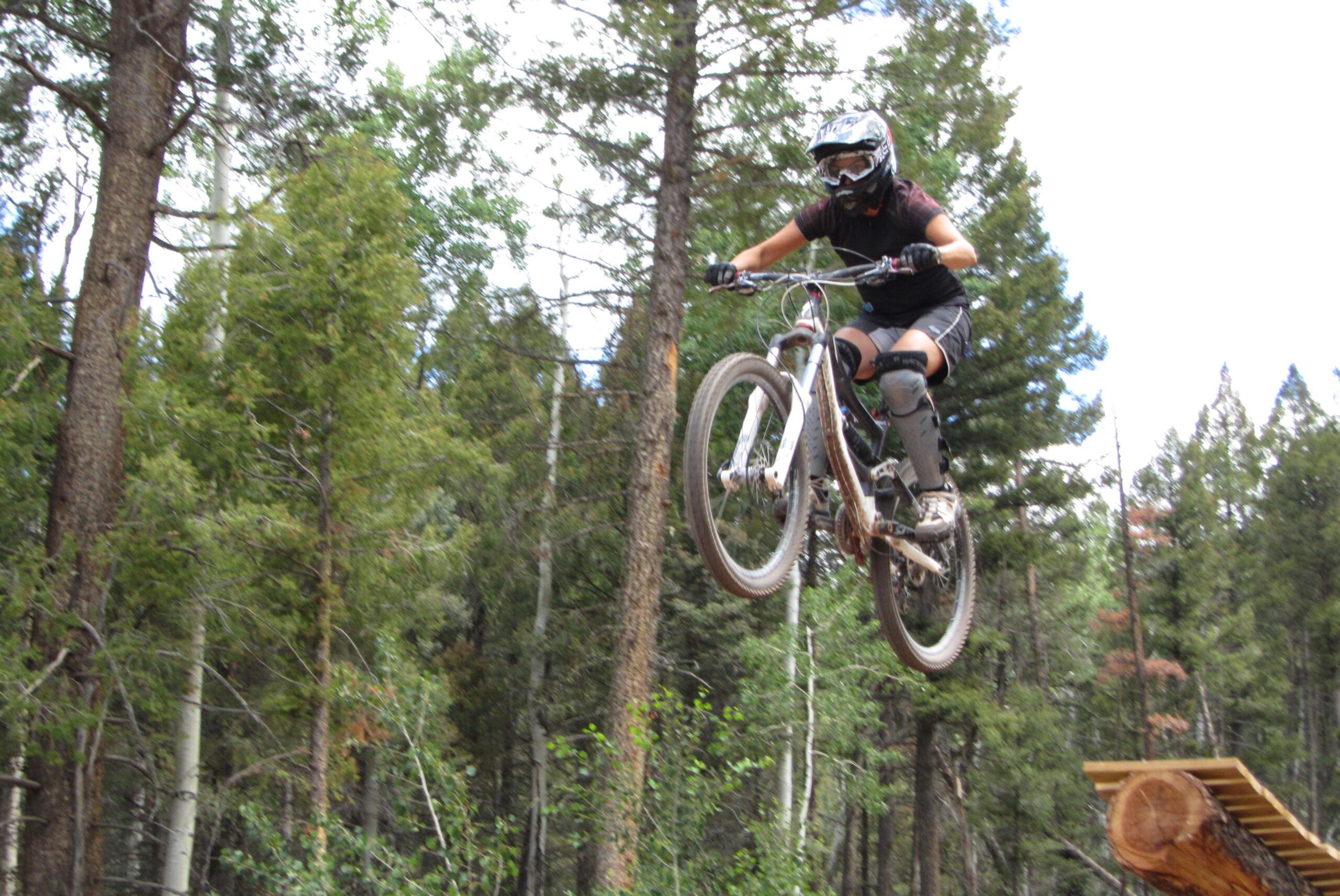 A mountain biker in protective gear jumps off a wooden ramp, soaring through the air with trees in the background. Angel Fire Bike Park mountain bike trail.