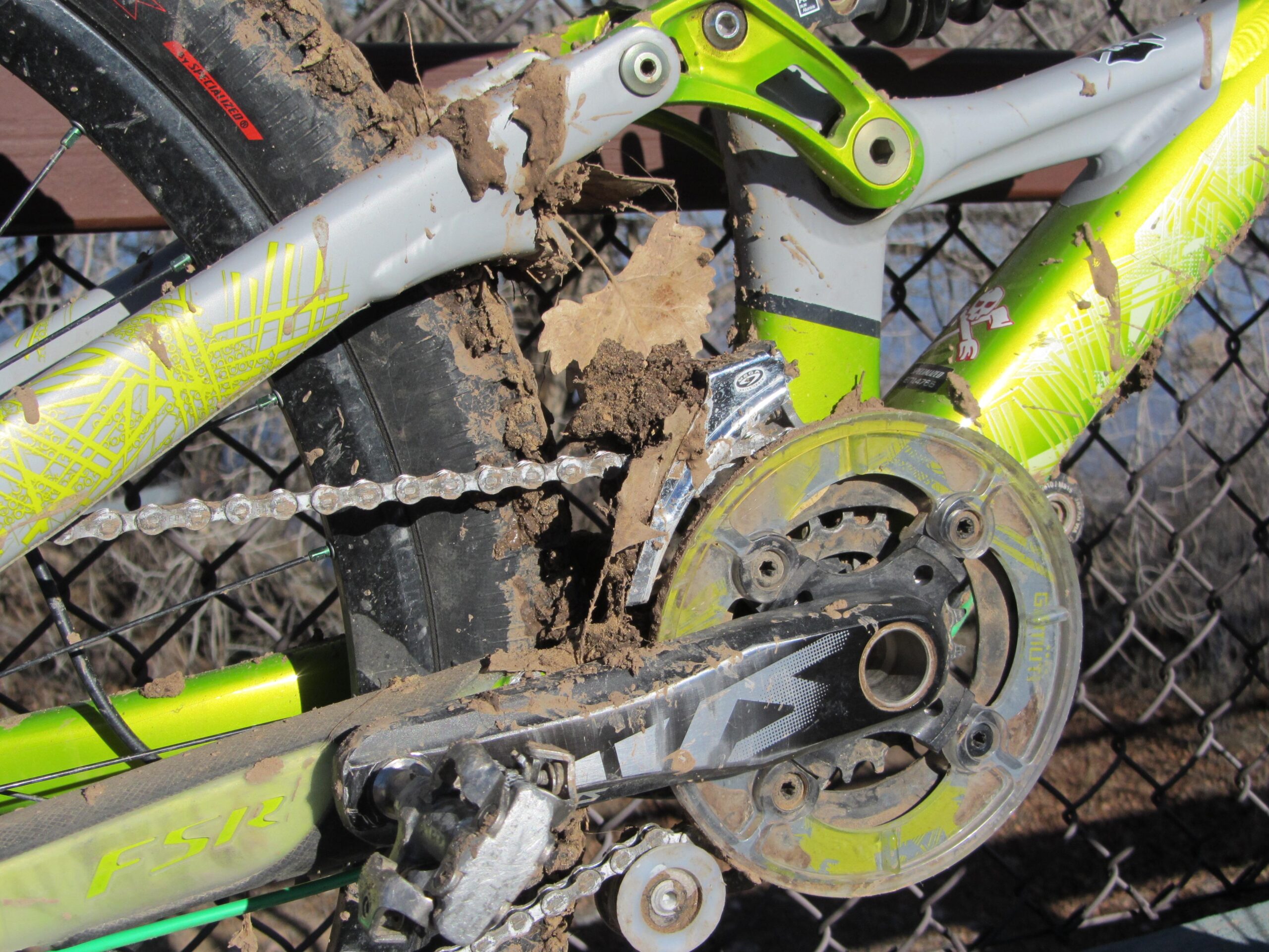 Close-up image of a mountain bike's drivetrain and frame, covered in mud and dirt after riding on a rugged trail. The bike features a vibrant green and silver design, with a visible chain and gears. A blurred background shows a fence and natural surroundings. Albuquerque Bosque mountain bike trail.