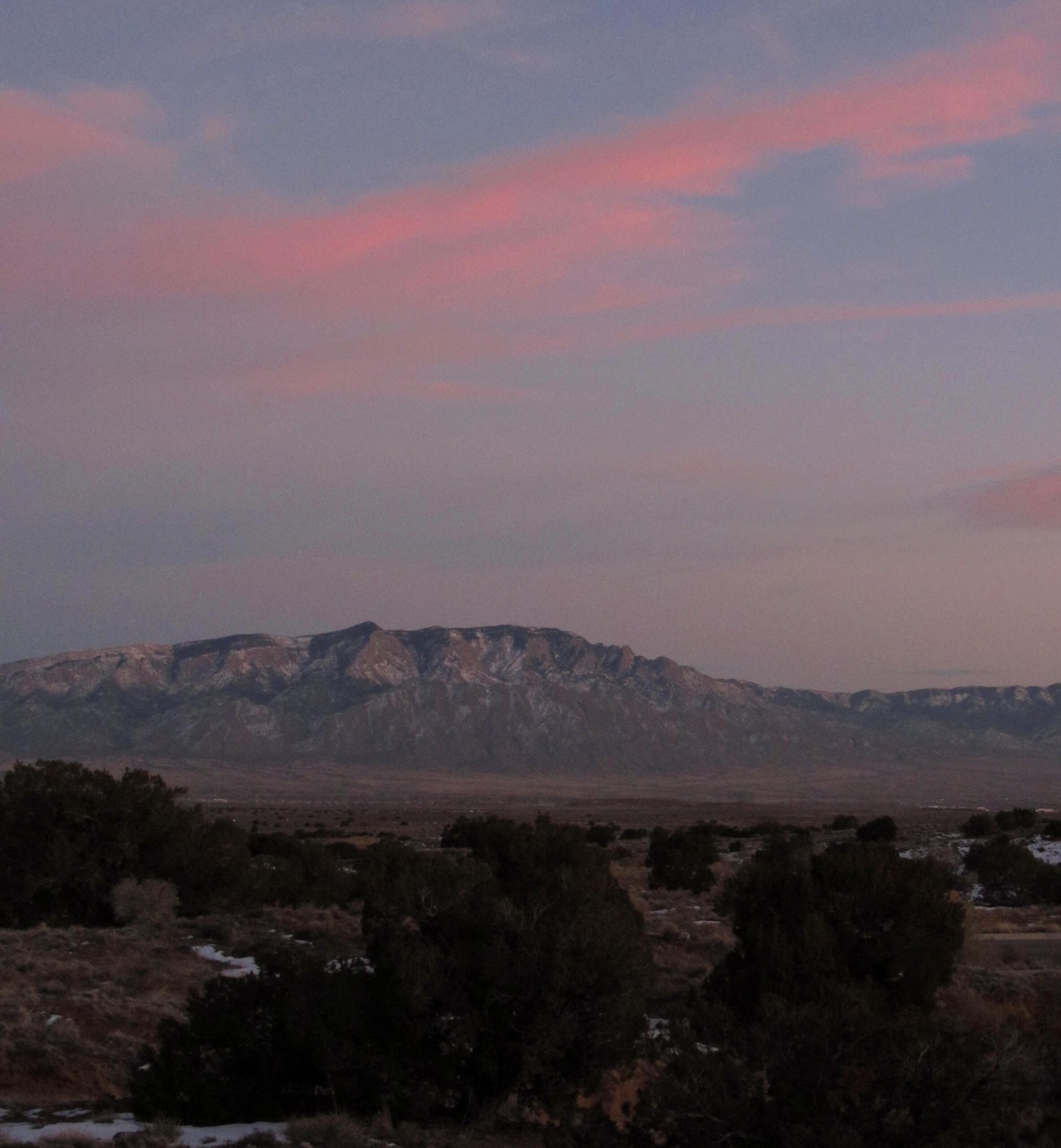A panoramic view of a mountainous landscape at dusk, featuring soft pink and purple hues in the sky. The foreground includes sparse vegetation and patches of snow, while the distant mountains are partially covered in snow, showcasing their rugged terrain. The overall atmosphere is serene and tranquil. Albuquerque Bosque mountain bike trail.
