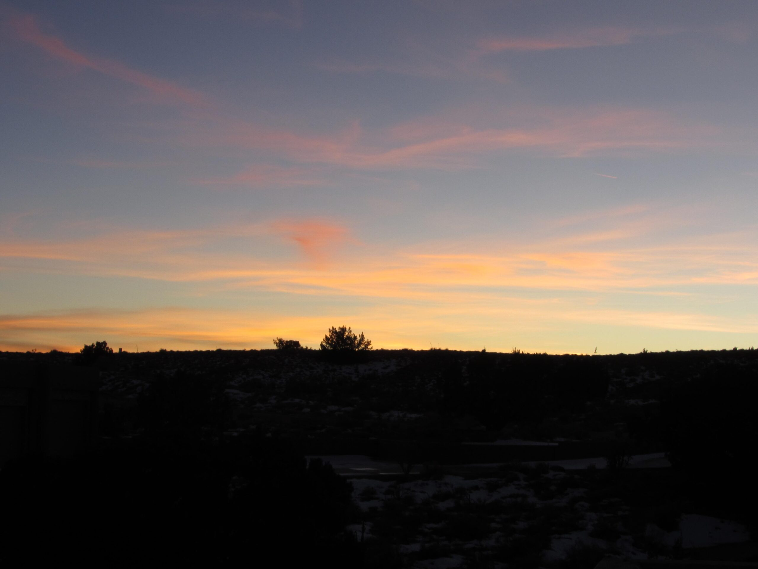 A serene sunset over a hilly landscape, featuring a gradient of colors from orange to pink in the sky, with dark silhouettes of trees and hills in the foreground. Albuquerque Bosque mountain bike trail.