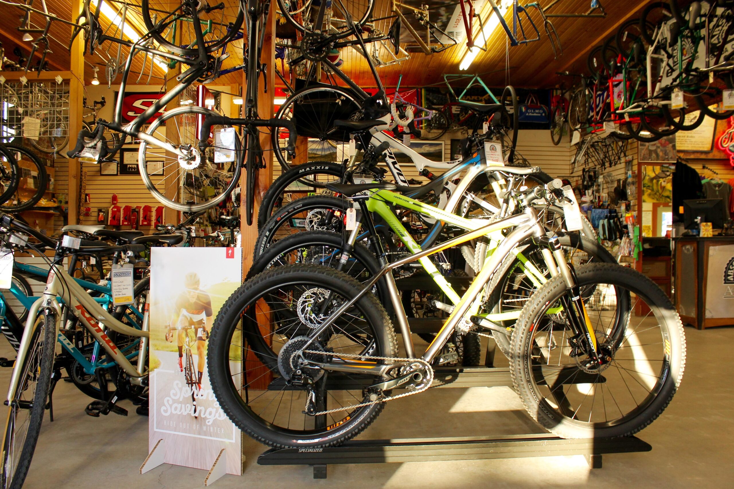 A vibrant bike shop interior displaying a variety of bicycles. Several bikes are prominently featured on a stand, with additional bikes hanging from the walls. A promotional sign for "Spring Savings" is visible in the foreground, indicating seasonal discounts. The shop has a warm ambiance with wooden accents and organized merchandise in the background.