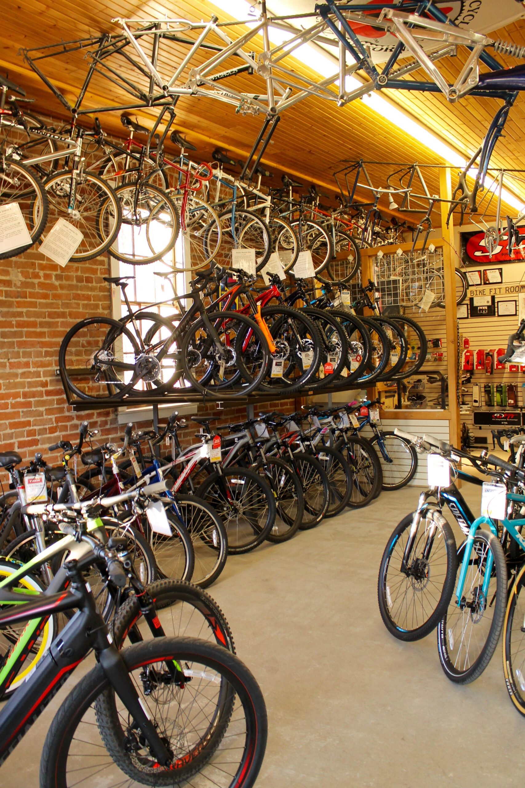 A bicycle shop interior featuring various bicycles displayed on racks. The walls are adorned with hanging bike frames, while a variety of multi-colored bicycles are positioned on shelves and the floor. Informational tags are visible on some bikes, and a brick wall adds to the shop's ambiance.