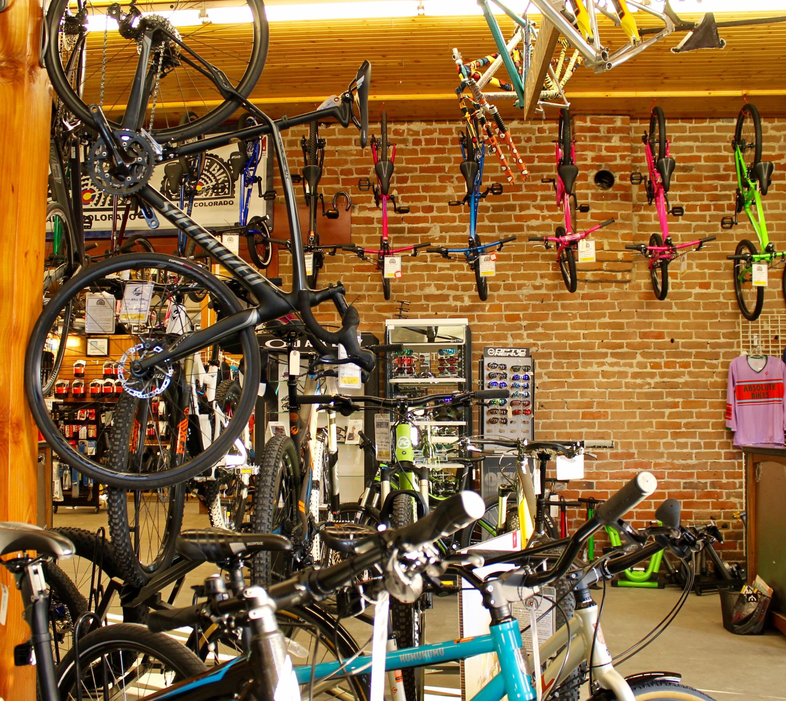 Inside a bicycle shop featuring a variety of bikes, both displayed on the walls and arranged on the floor. There are different styles of bicycles, including road and mountain bikes, along with colorful BMX bikes hanging from the ceiling. The interior has a rustic feel with exposed brick walls and wooden beams, while various cycling accessories are visible in the background.