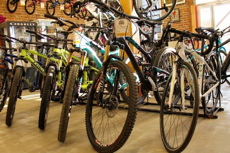 A display of various bicycles in a bike shop, featuring mountain bikes in green and blue colors in the foreground, with a sleek road bike in white to the right. The scene includes a backdrop of brick walls and multiple bicycles hanging from the ceiling.