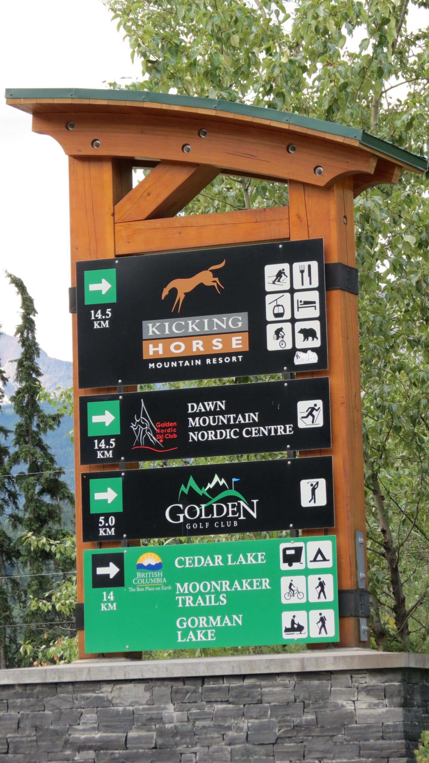 Wooden directional sign for outdoor activities at Kicking Horse Mountain Resort, featuring distances to various locations including Kicking Horse, Dawn Mountain Nordic Centre, Golden Golf Club, Cedar Lake, Moonraker Trails, and Gorman Lake. The sign includes icons for skiing, dining, and other amenities, set against a backdrop of trees and mountains. Moonraker Trails mountain bike trail.