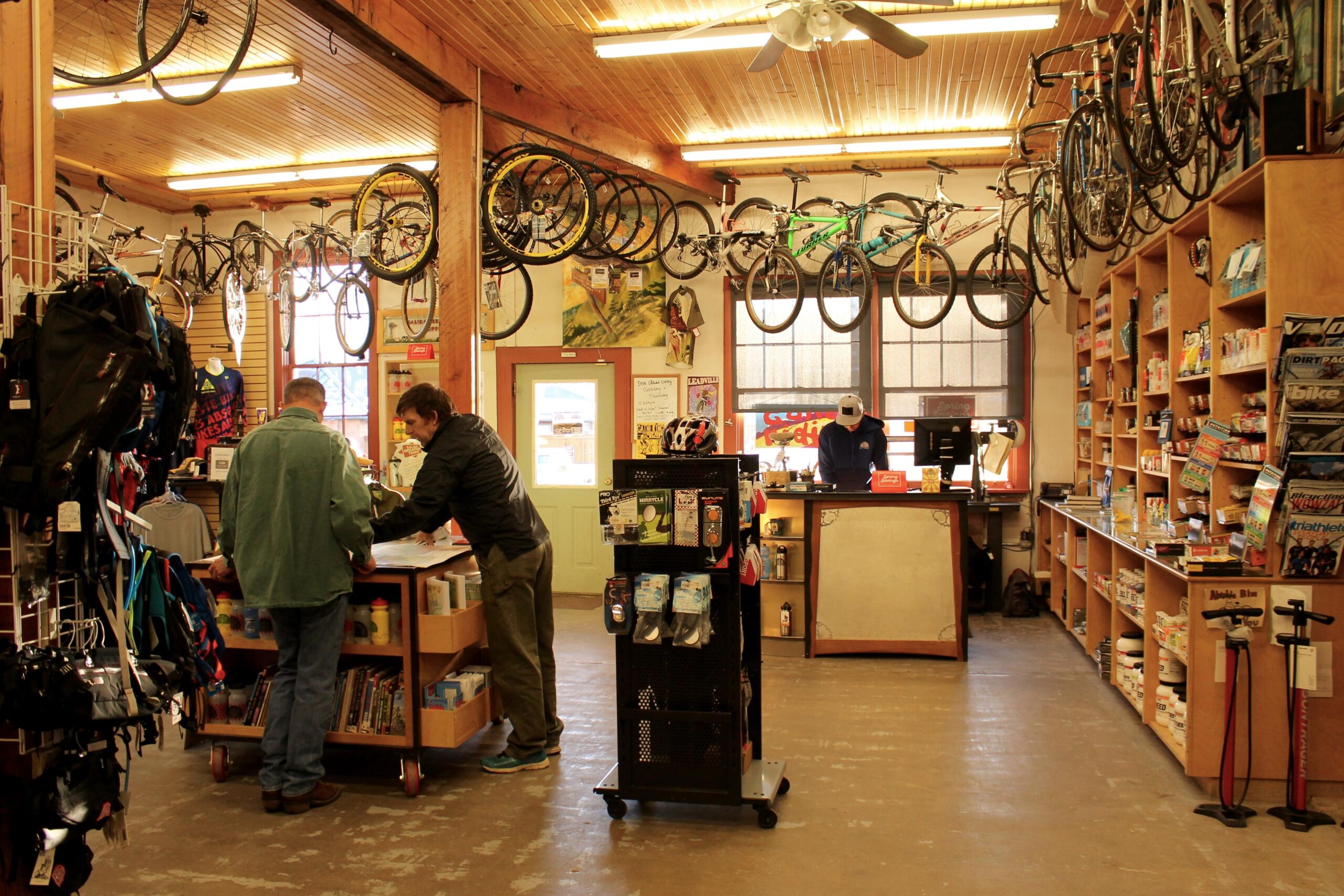 A bike shop interior featuring two customers examining products at a central display table. The background is filled with various bicycles hanging from the ceiling and shelves stocked with cycling gear, accessories, and magazines. A sales associate is visible behind the counter, attending to a transaction. The space has a warm, inviting atmosphere with wood paneling and bright lighting.