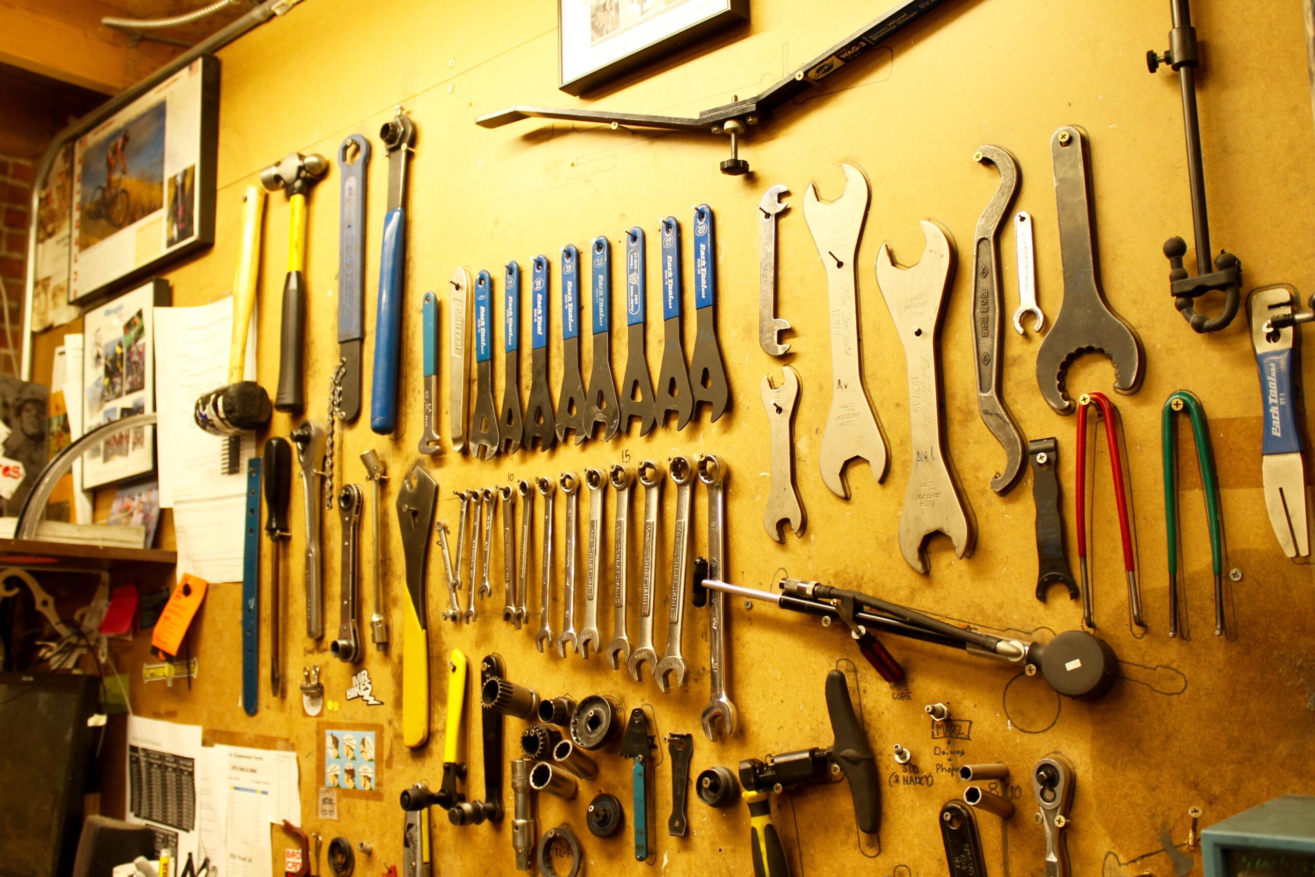 A wall filled with various hand tools, including wrenches, a mallet, and other mechanical equipment, organized on a wooden surface. There are also sheets of paper and photographs displayed above the tools. The scene captures a workshop or garage setting.