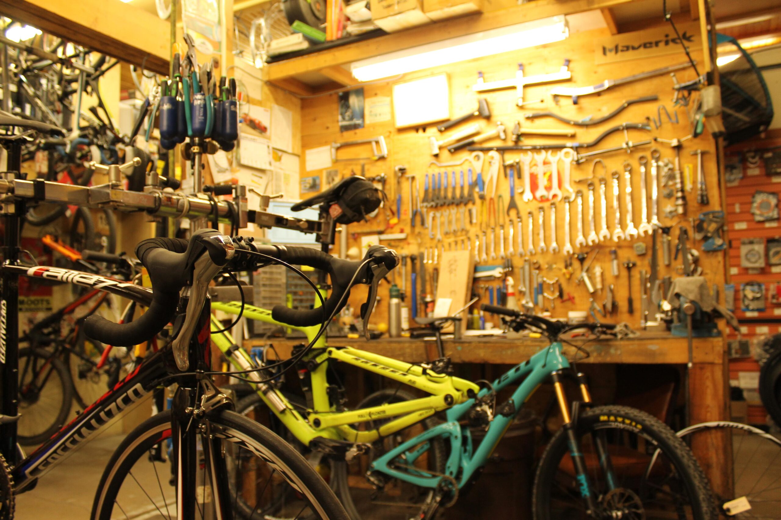 A busy bike workshop filled with various bicycles and tools. In the foreground, a sleek black road bike is prominently displayed, while a bright green mountain bike is partially visible in the background. The wall behind is lined with an array of tools, including wrenches, screwdrivers, and other bike repair equipment, creating a vibrant and functional atmosphere for bike maintenance.