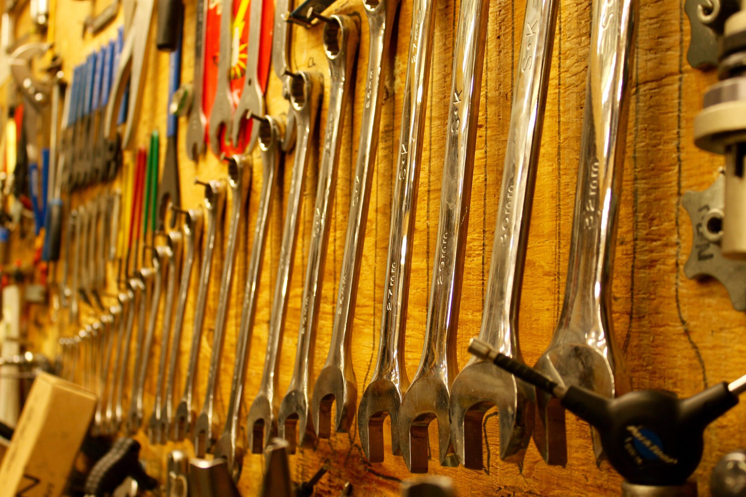 A close-up image of a wall-mounted display featuring an array of shiny, metallic wrenches arranged in a row. The background consists of a wooden surface, and various other tools are visible, including pliers and screwdrivers, creating a workshop or garage ambiance.