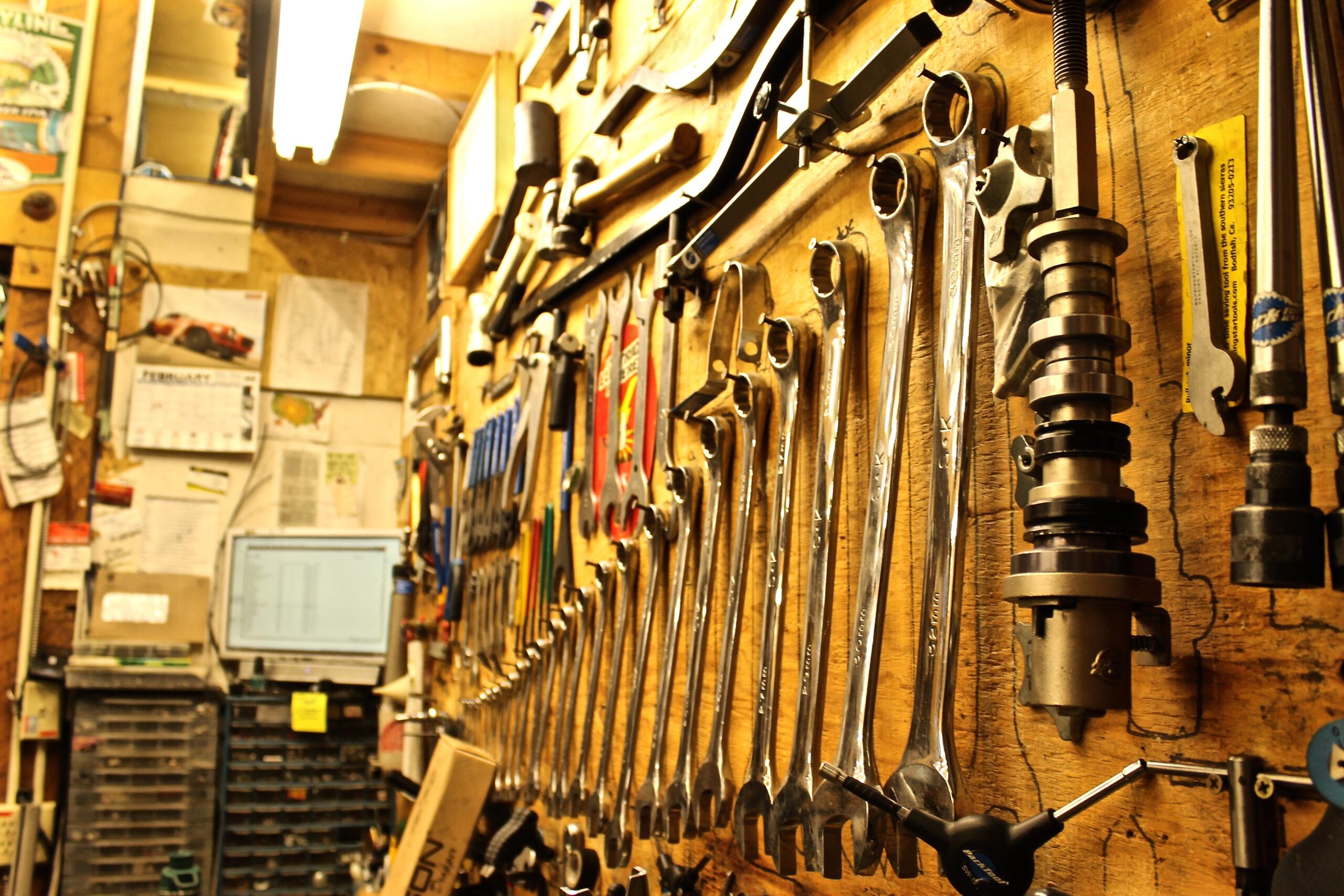 A well-organized workshop wall featuring an array of metallic tools, including wrenches and a camshaft, hanging neatly on a wooden backdrop. In the background, there are scattered papers, a calendar, and a computer monitor displaying a spreadsheet. The scene exudes a practical and industrious atmosphere.