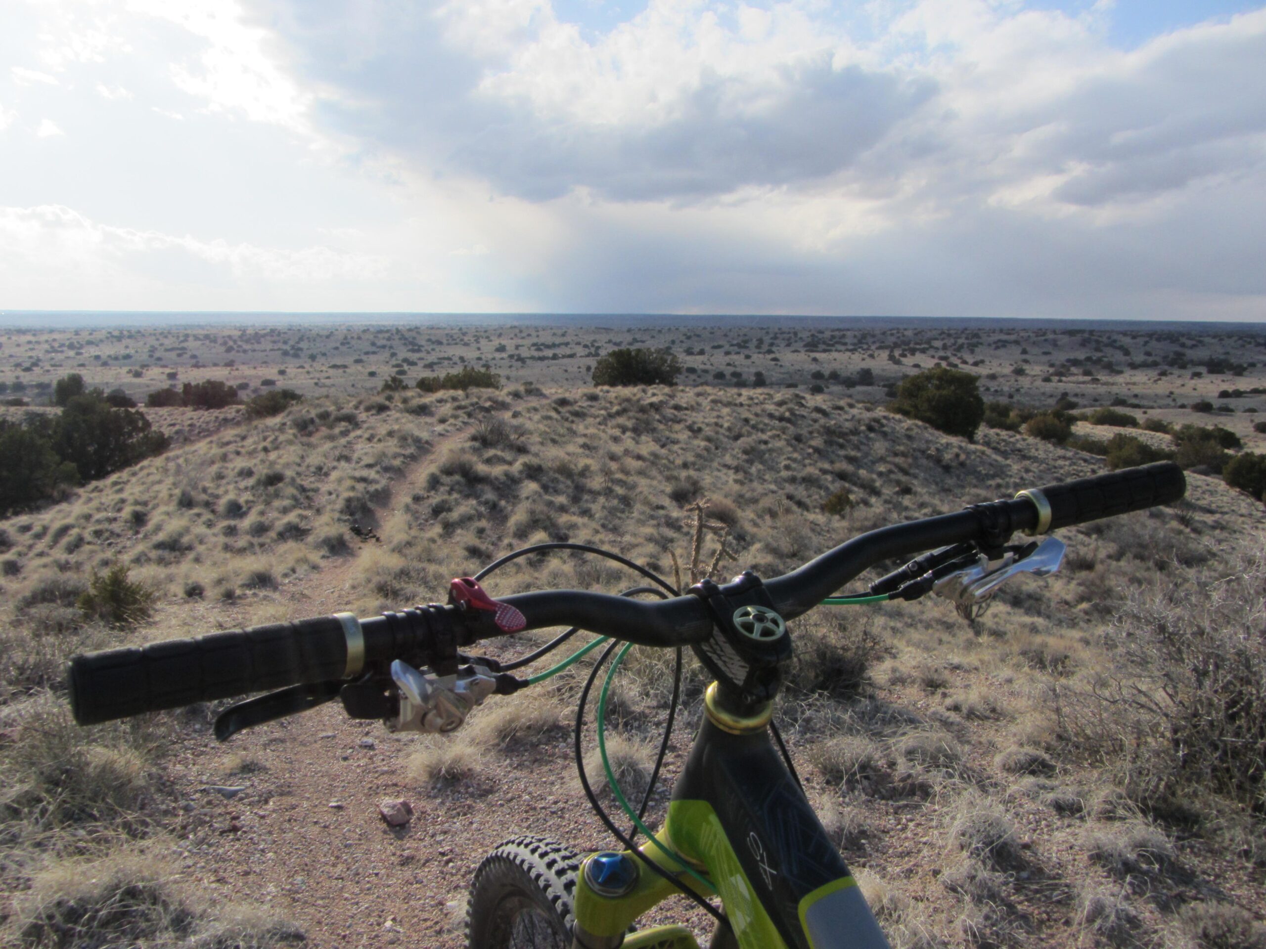 Mountain bike handlebars in the foreground, overlooking a vast, open landscape with sparse vegetation and rolling hills under a cloudy sky. Reservoir Fatbike trail mountain bike trail.
