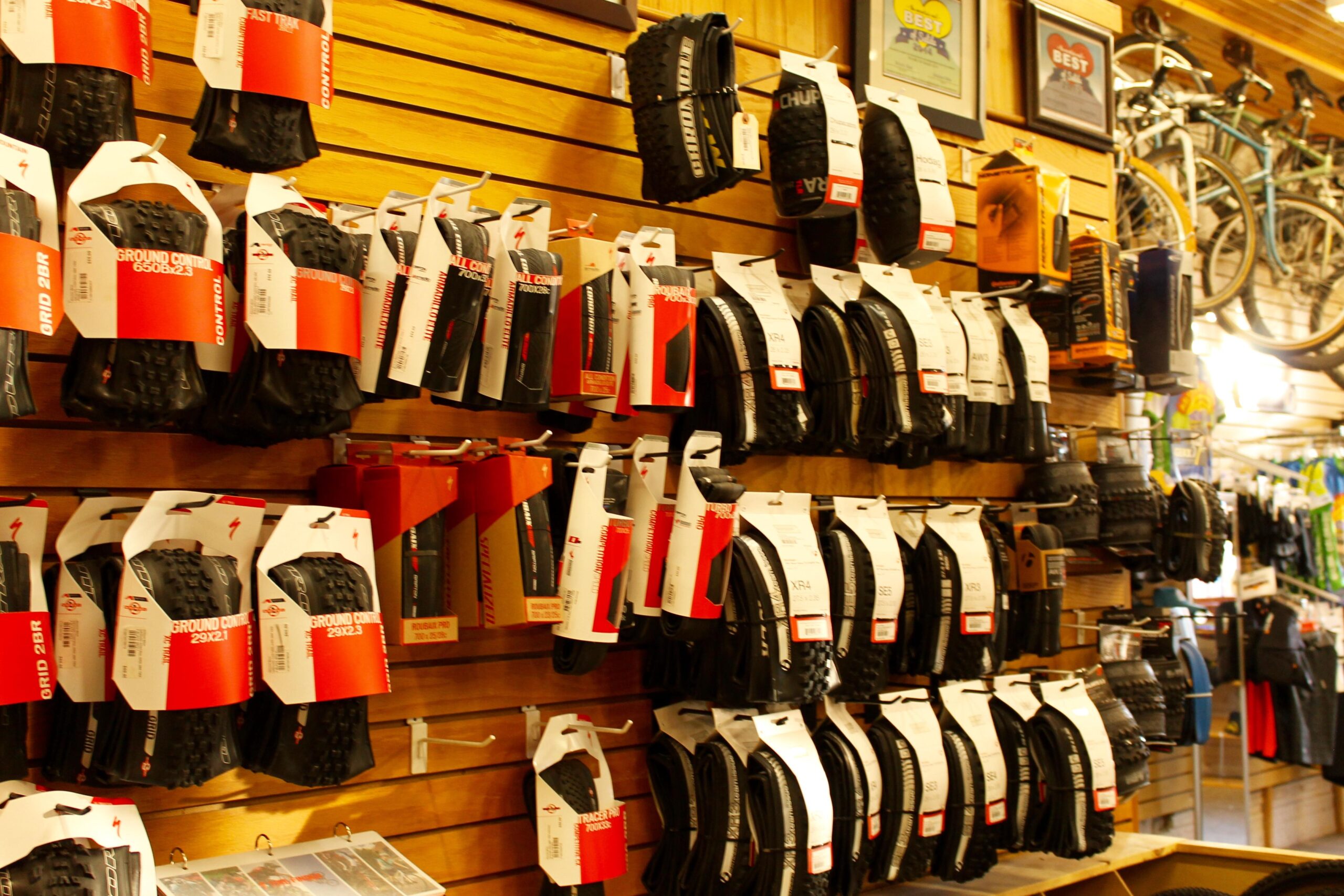 A display of various bicycle tires arranged on wooden shelves in a bike shop, featuring multiple brands and styles, with packaging labels visible. In the background, bicycles and other cycling accessories are partially visible.