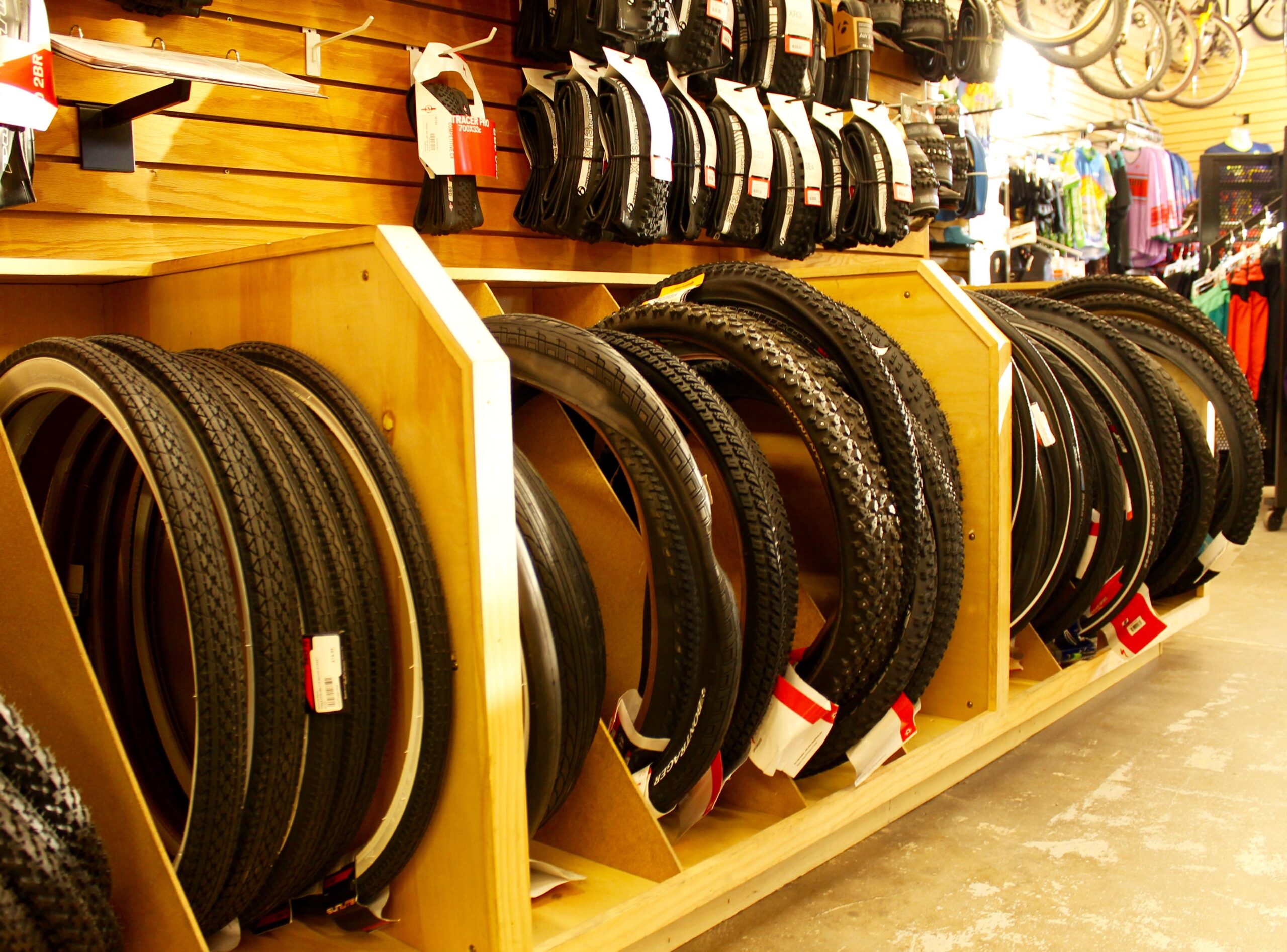 Image of a bicycle tire display in a shop, featuring various types of tires neatly arranged on wooden shelves. The background includes additional bike accessories and clothing for sale.