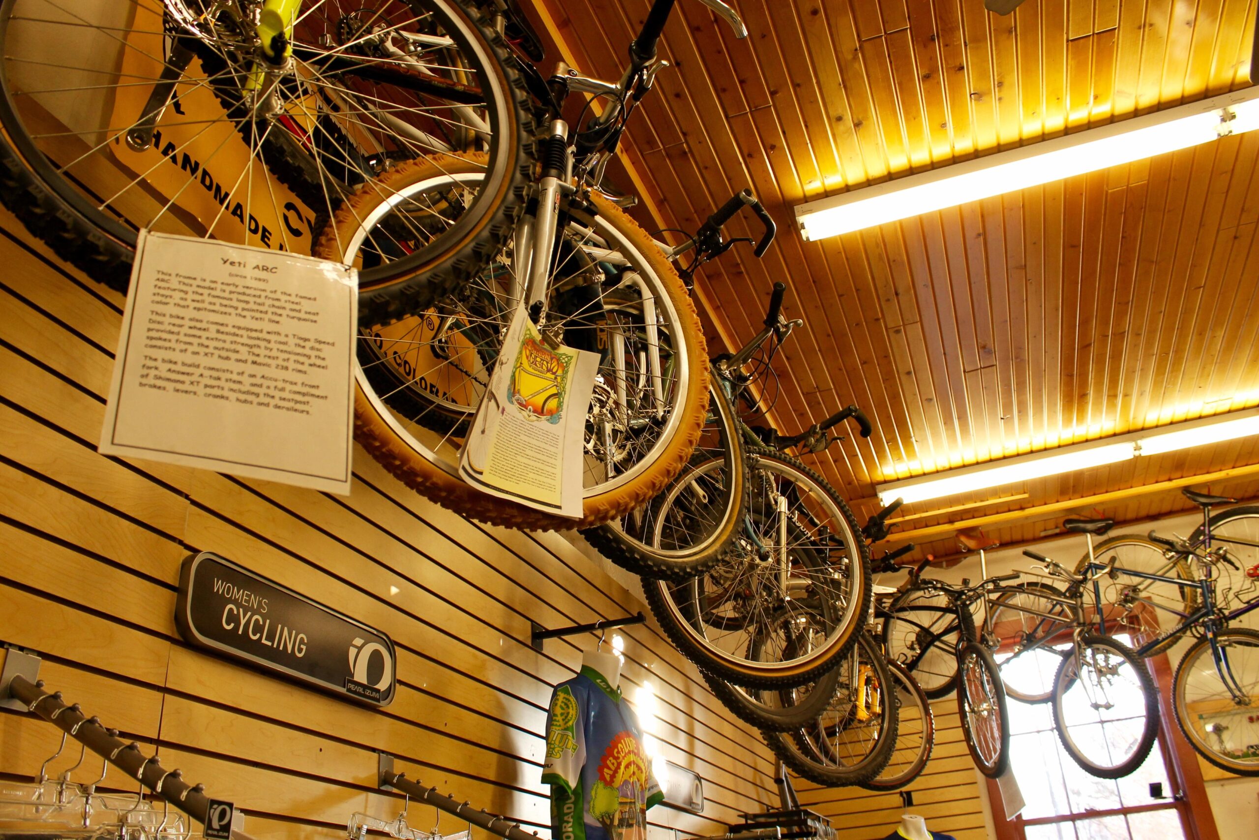A display of bicycles hanging from wooden panels in a bike shop, featuring various models and a sign labeled "Women's Cycling." A detailed information card about the Yeti ARC bike is visible, along with colorful cycling jerseys hanging below. Bright overhead lighting illuminates the scene.