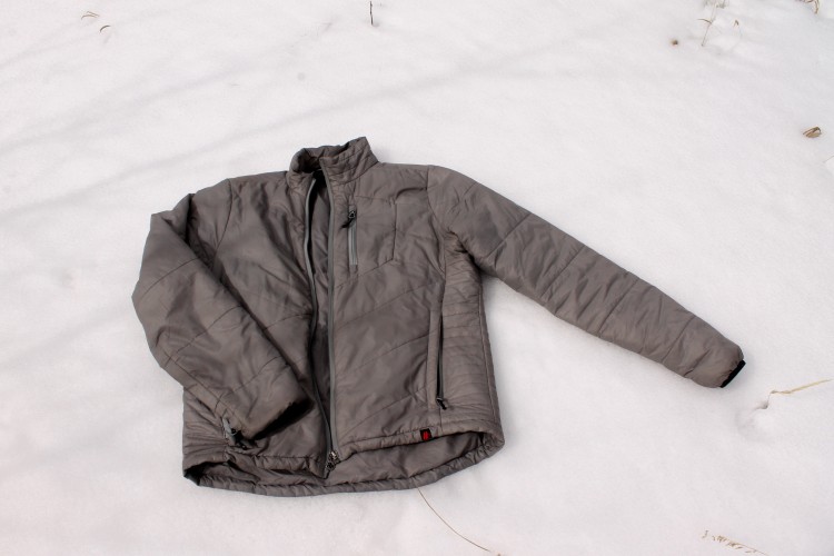 A gray insulated jacket lying on a snowy ground with some grass peeking through the snow.